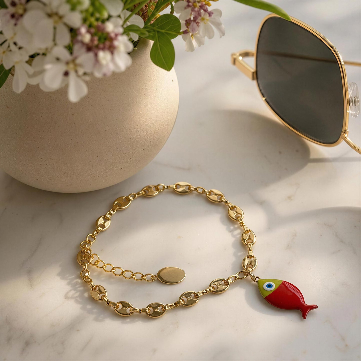 Gold bracelet with a fish charm on a marble surface next to a vase of flowers and sunglasses.