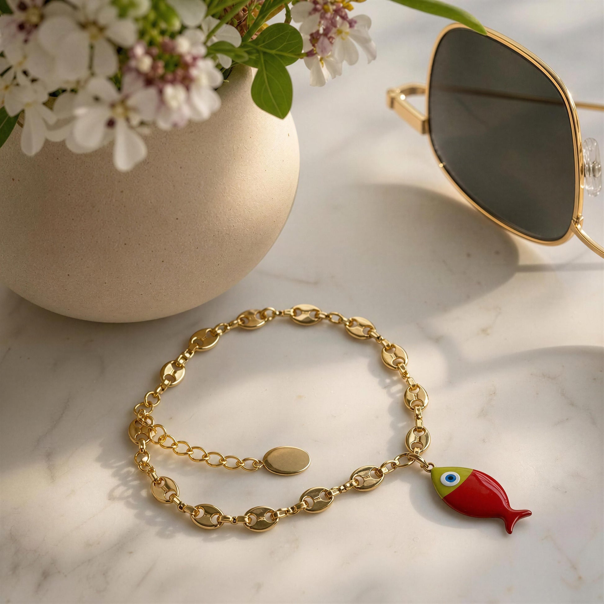 Gold bracelet with a fish charm on a marble surface next to a vase of flowers and sunglasses.