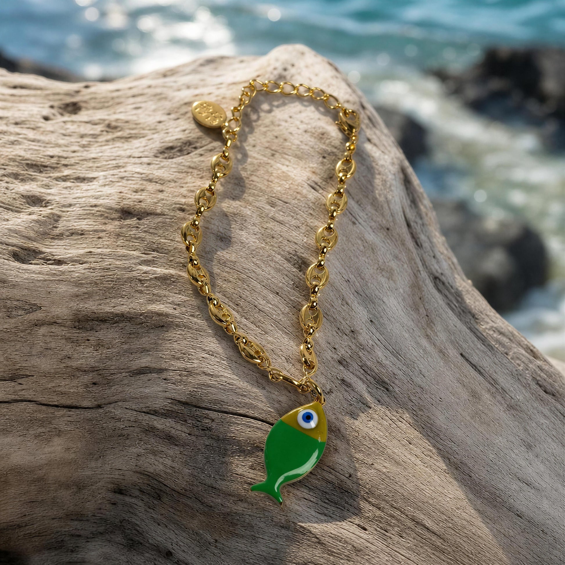 Gold bracelet with a green fish charm on a piece of driftwood by the water.