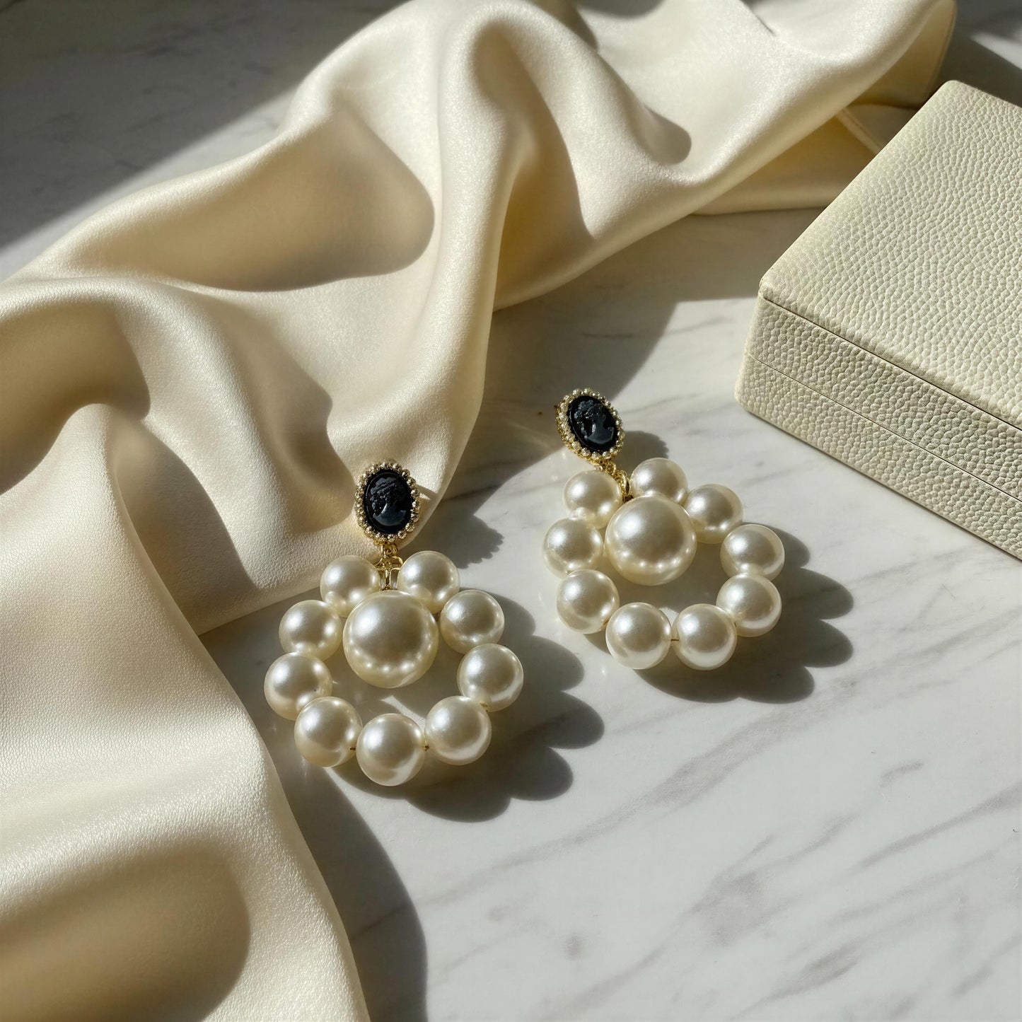 The Regal Cameo Pearl Statement Earrings on a marble surface with fabric and jewelry box.