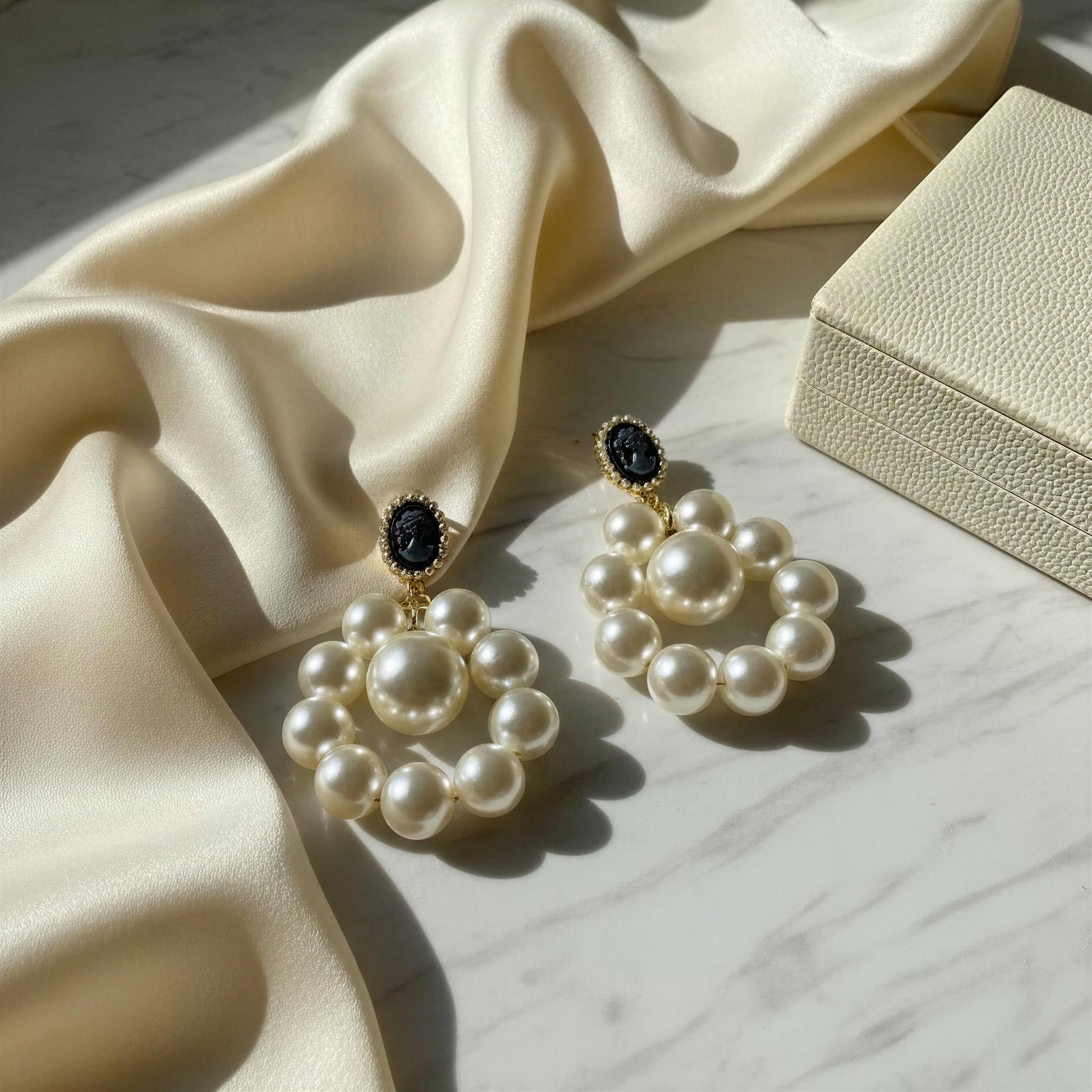 The Regal Cameo Pearl Statement Earrings on a marble surface with fabric and jewelry box.