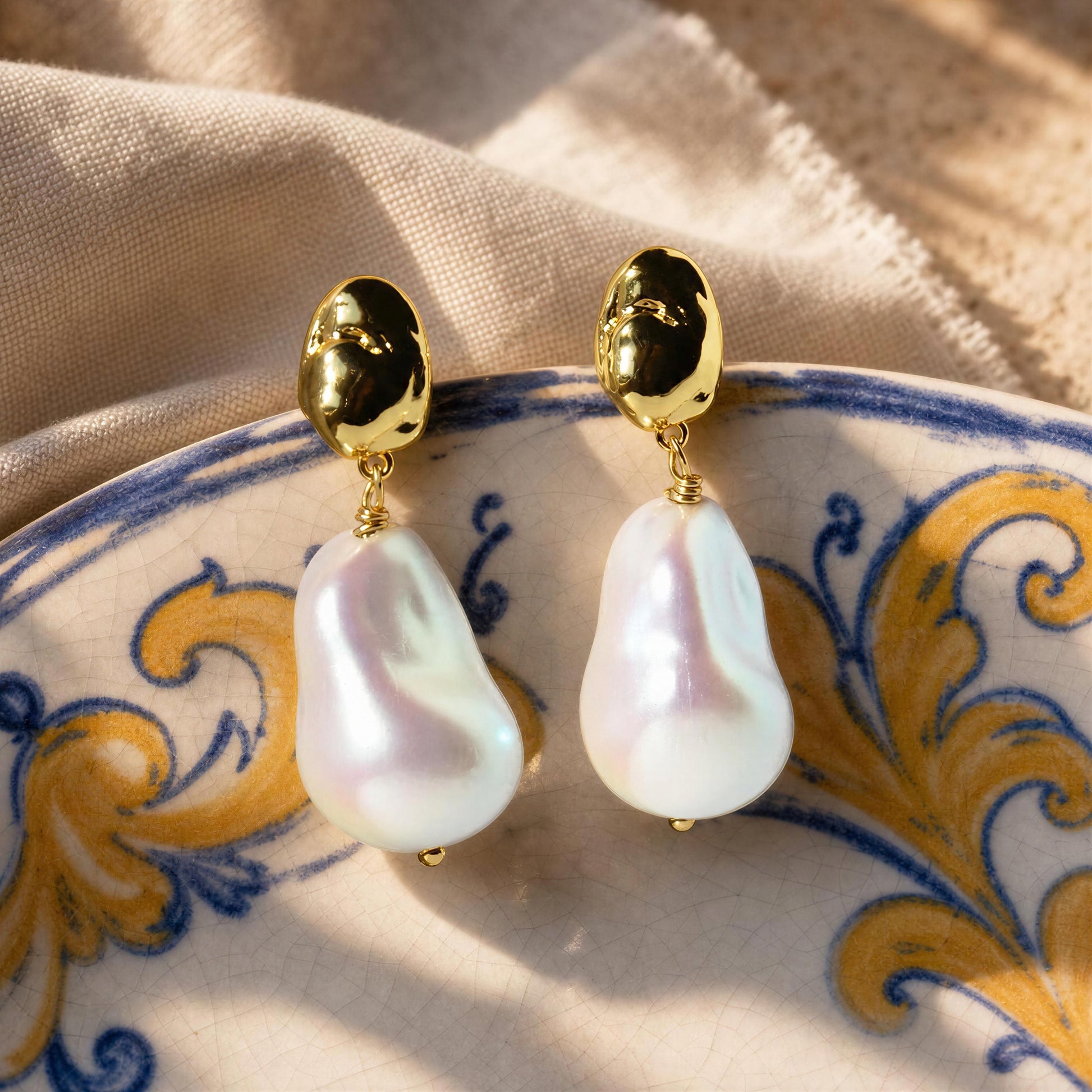 A pair of gold drop earrings with large, irregular white baroque pearls, displayed on a decorative blue and gold patterned ceramic plate over a beige fabric background.
