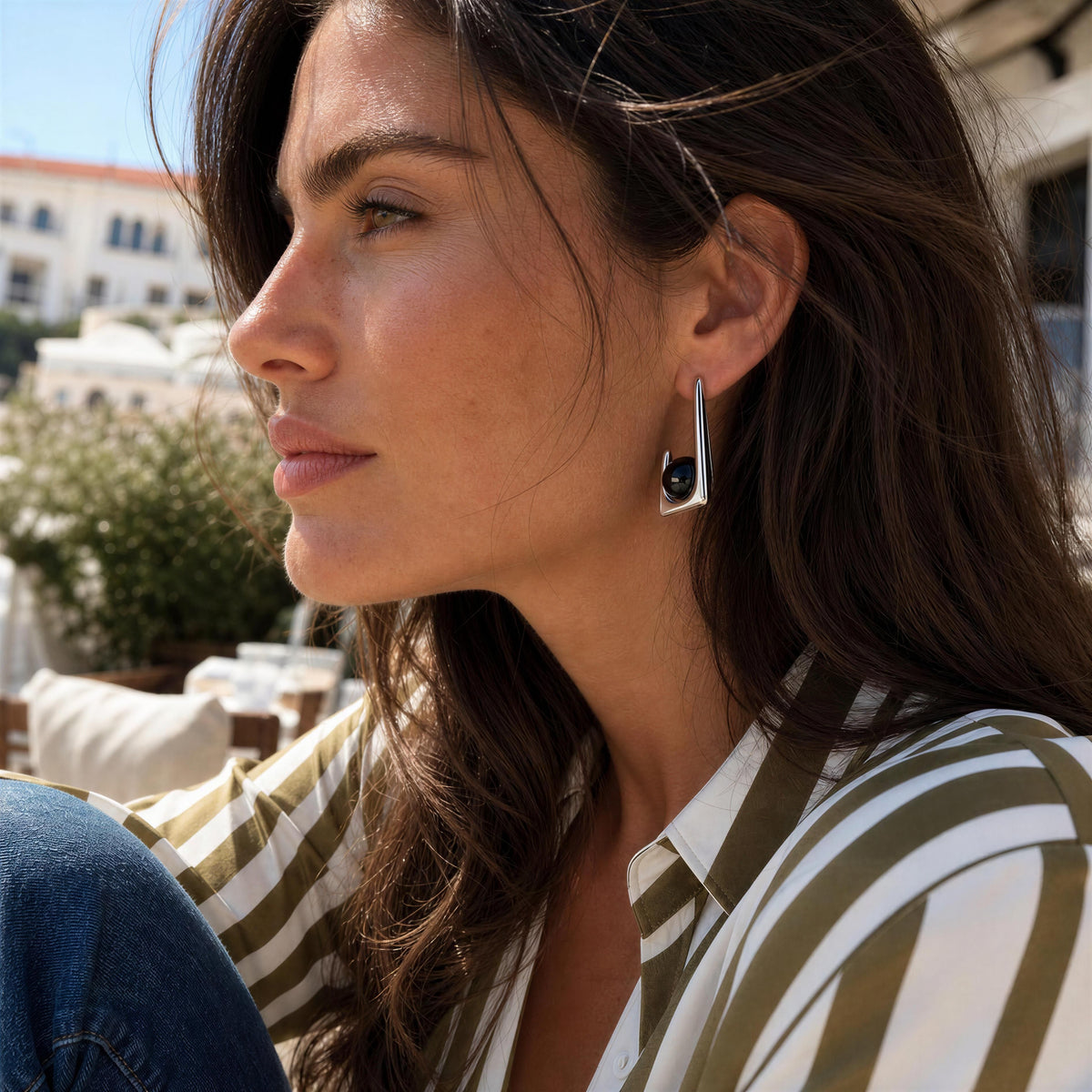 A woman in a striped shirt wearing the Chica_Chica_Jewelry Kara Earrings, silver geometric dangle earring with a black circular accent, photographed outdoors in bright natural light.