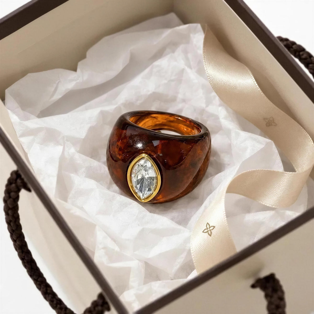 A chunky amber-brown resin ring with a marquise-cut clear gemstone in a gold bezel setting, presented inside a cream-colored gift box with white tissue paper and a satin ribbon.
