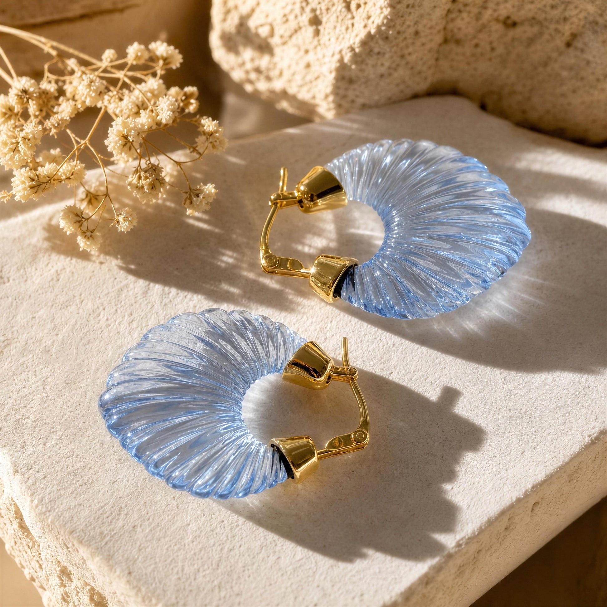 The Chica Chica Jewelry Celeste Earrings. A pair of light blue ribbed hoop earrings with gold-toned posts, displayed on a textured beige stone surface with dried flowers in the background.