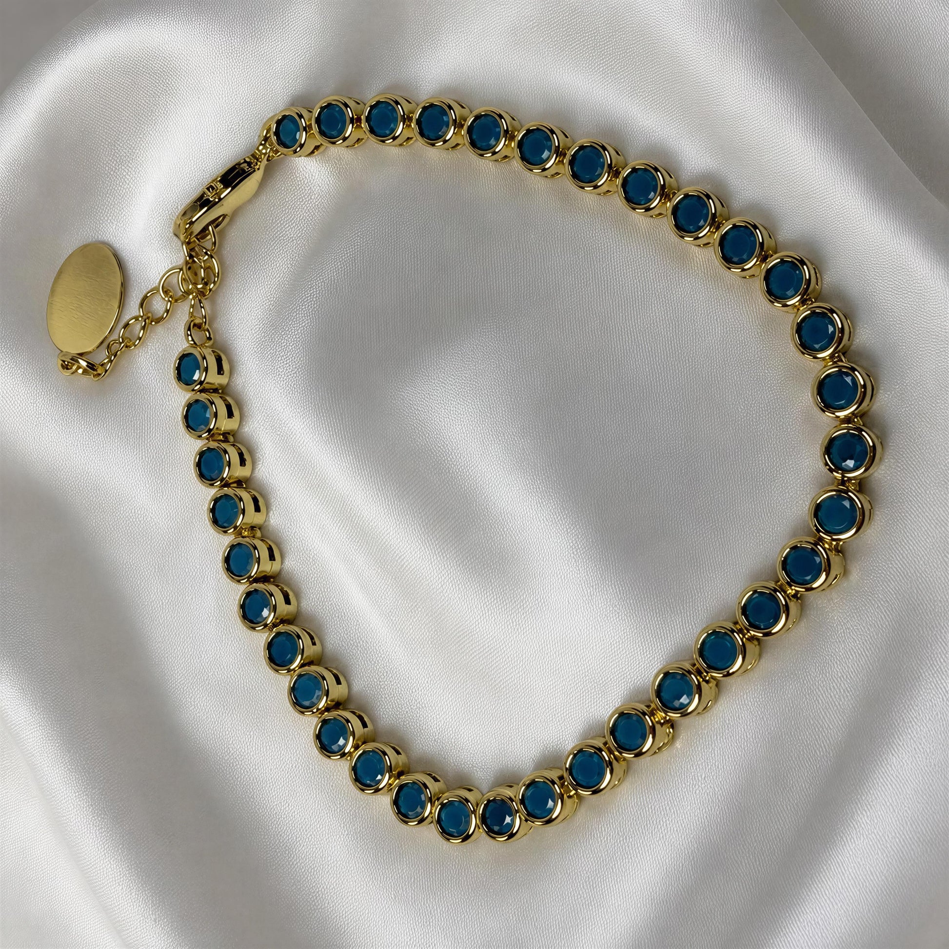 A gold-tone tennis-style bracelet with round turquoise gemstones, displayed on a white satin fabric background.