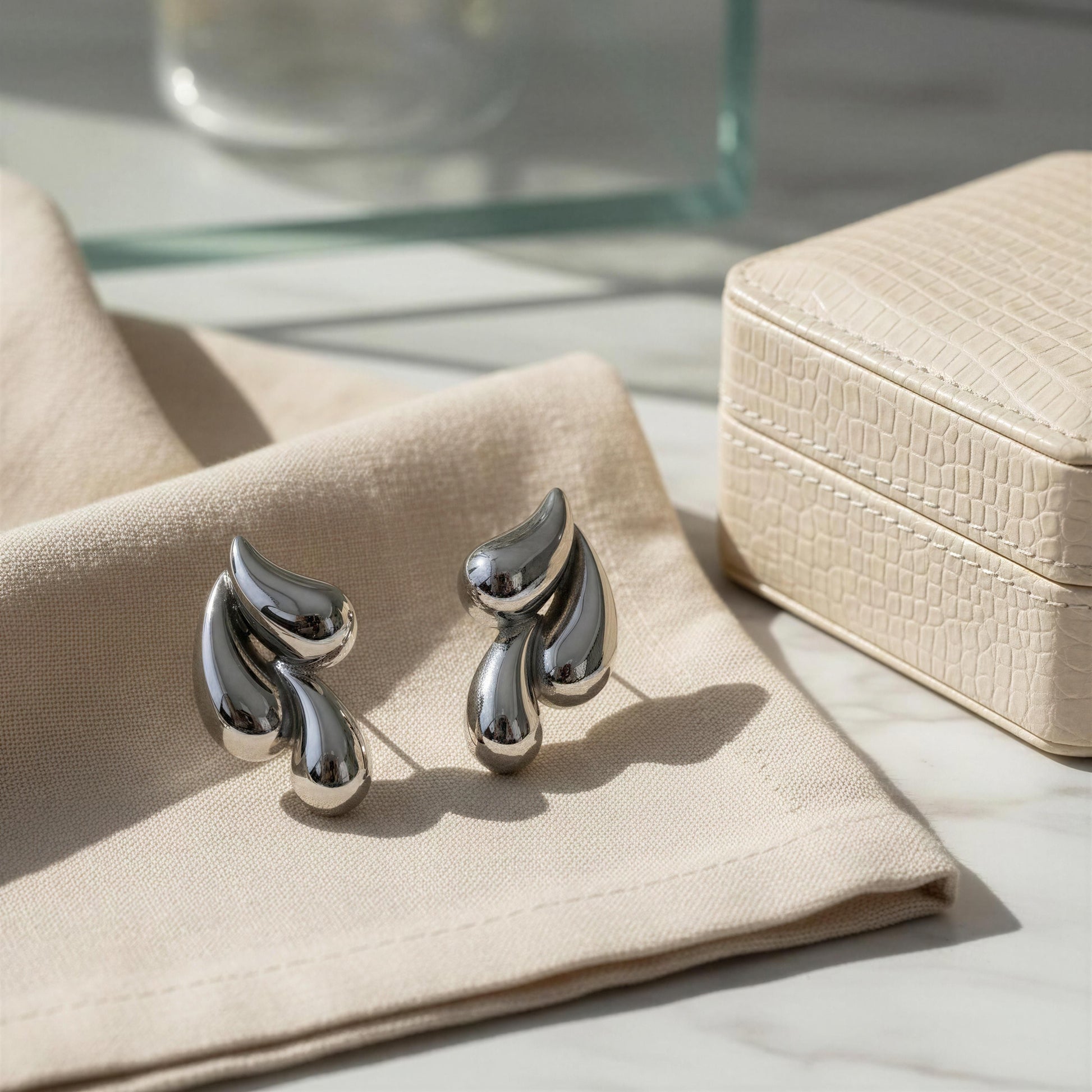 The Silver Molten Wave Drop Earrings on a beige cloth with a jewelry box in the background