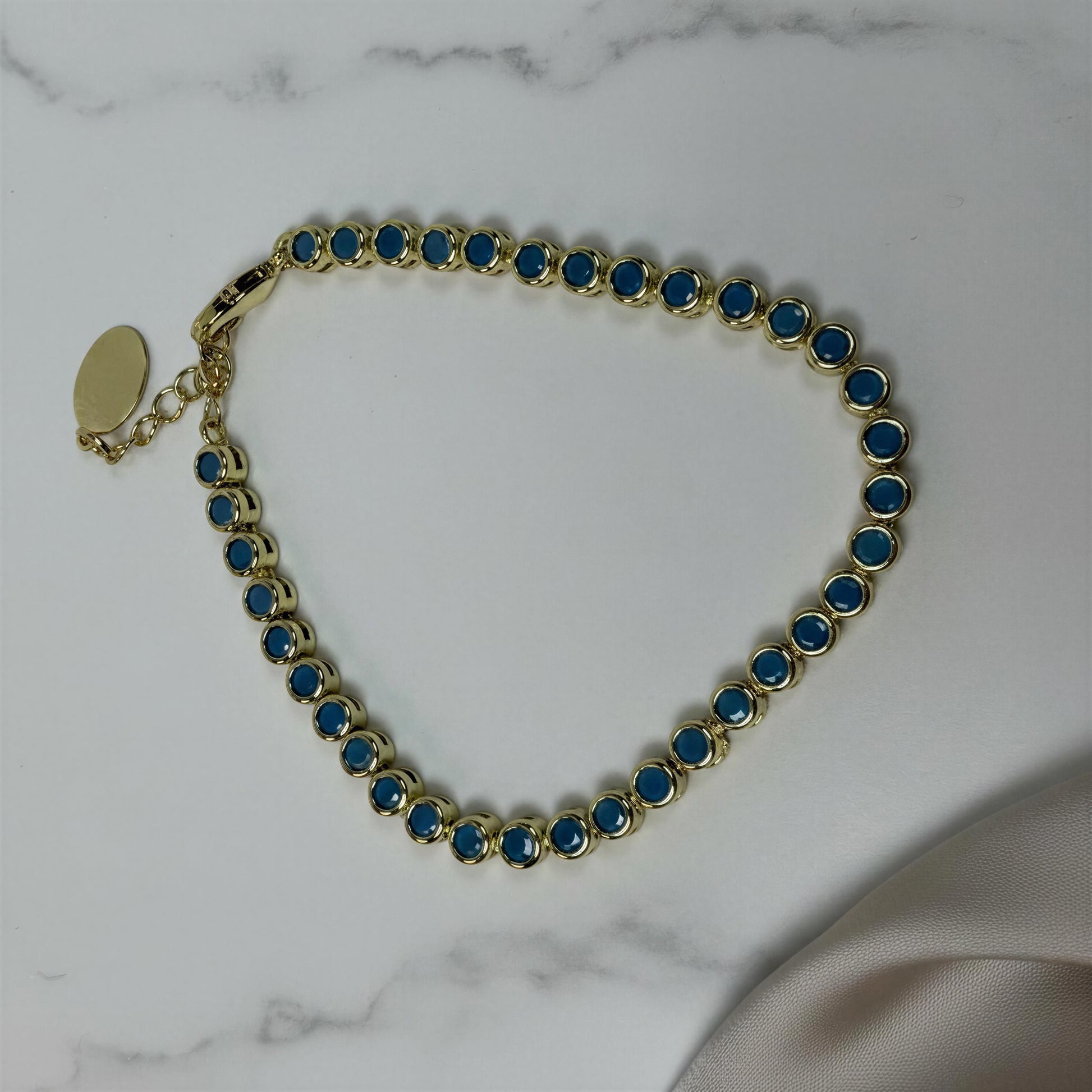 A gold tennis bracelet with round turquoise gemstones, displayed on a white marble surface with a beige satin fabric corner visible in the lower right.