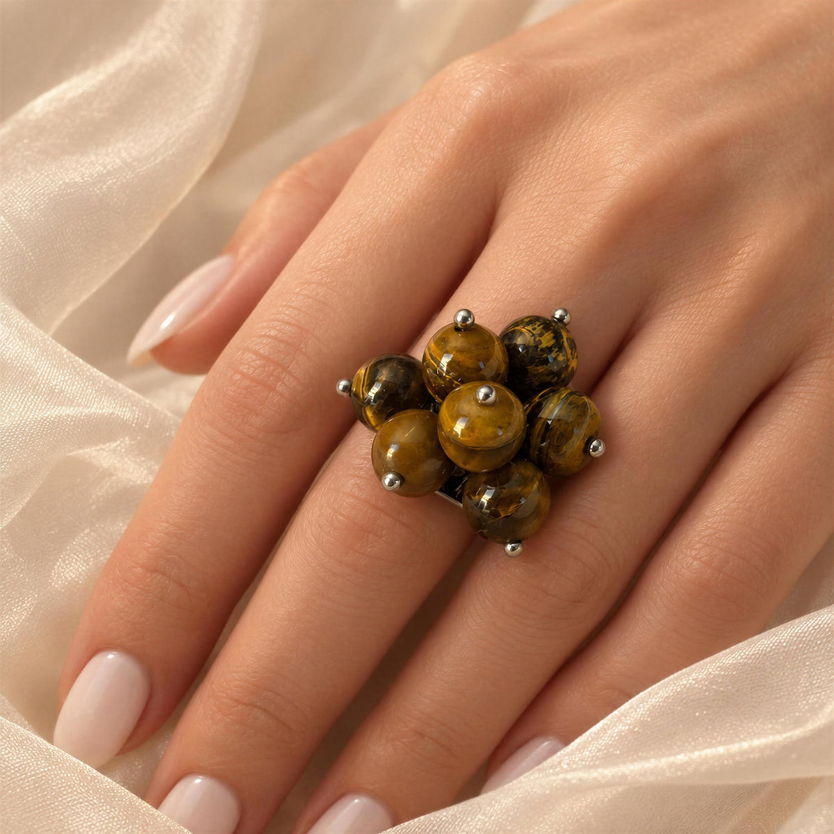 A close-up of a hand wearing the Talia Ring, a chunky ring made of clustered round tiger's eye beads with small silver prongs, set against soft cream-colored fabric.