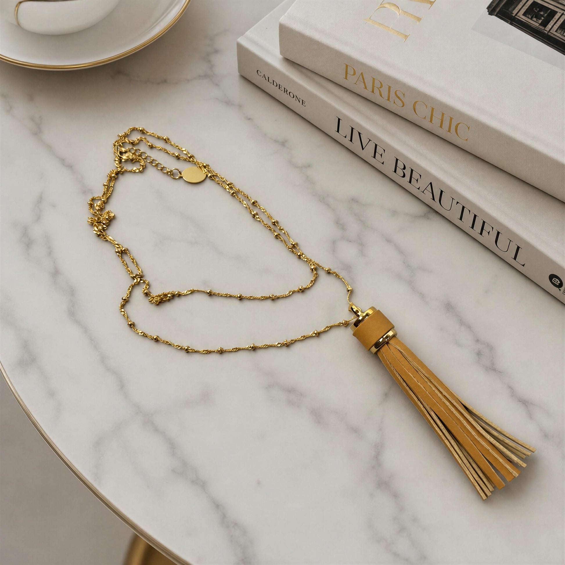 The Frangia Cammello tassel necklace featuring a camel faux leather tassel on a delicate 18K gold plated satellite chain, styled on a white marble tabletop next to stacked books.