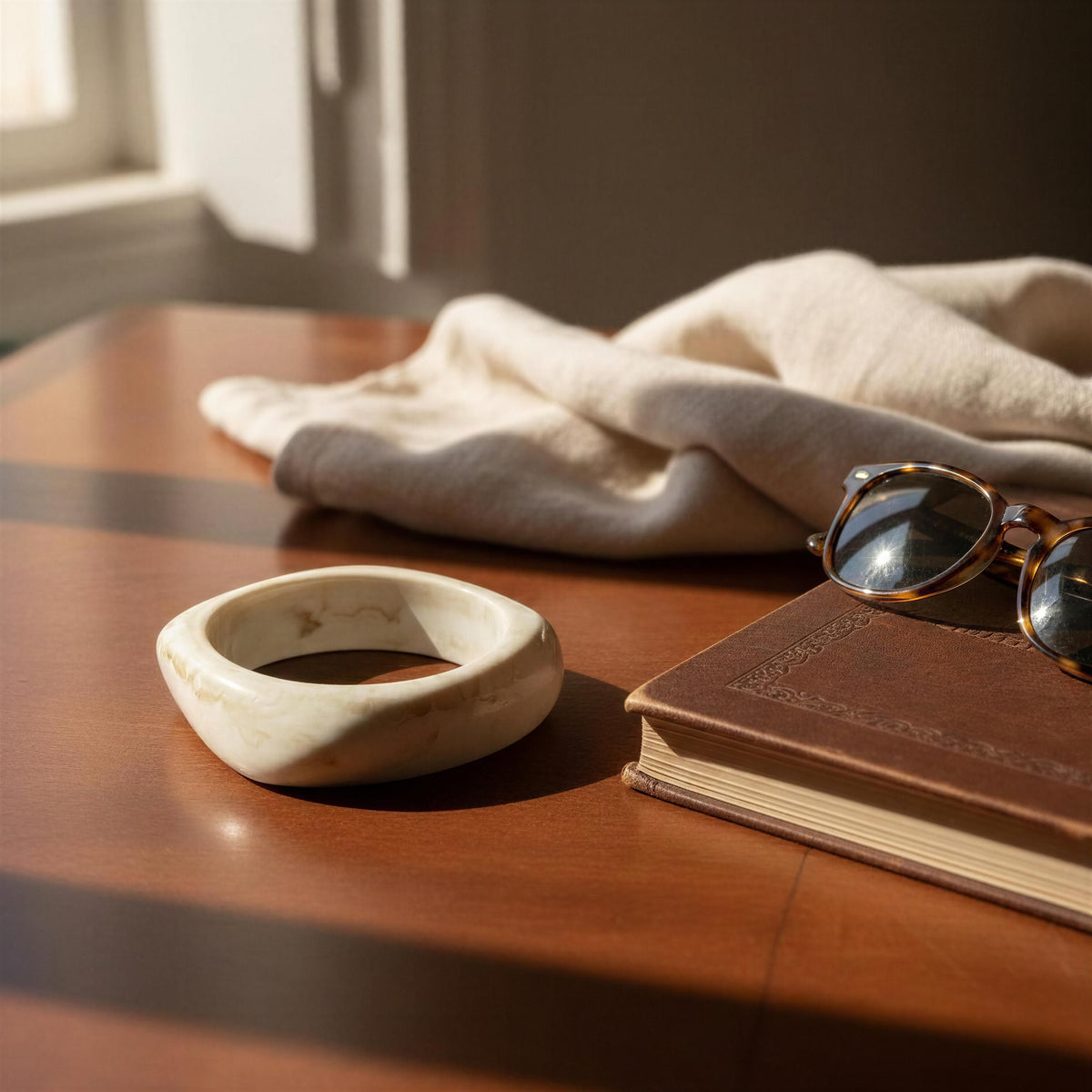A cream-colored, irregularly shaped stone bangle bracelet with subtle brown veining, displayed on a wooden table next to a brown leather book, tortoiseshell sunglasses, and a beige linen cloth in warm sunlight.