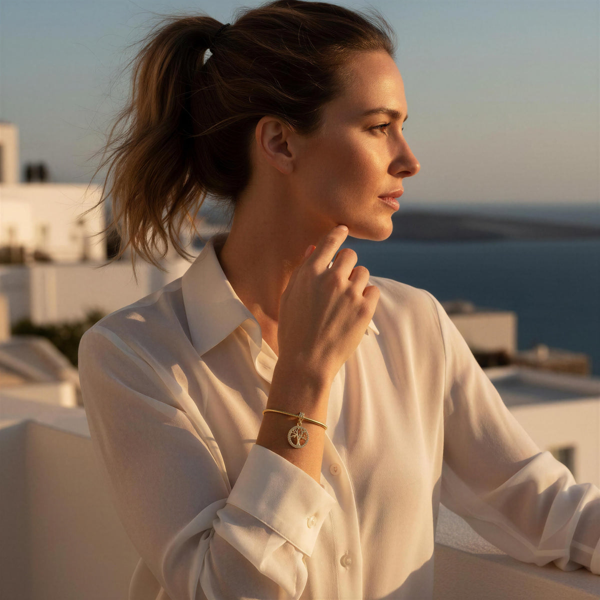 A woman in a white button-up shirt wearing a delicate gold chain bracelet with the tree of life charm, posing outdoors against a warm sunset background with a sea and white buildings in the distance.