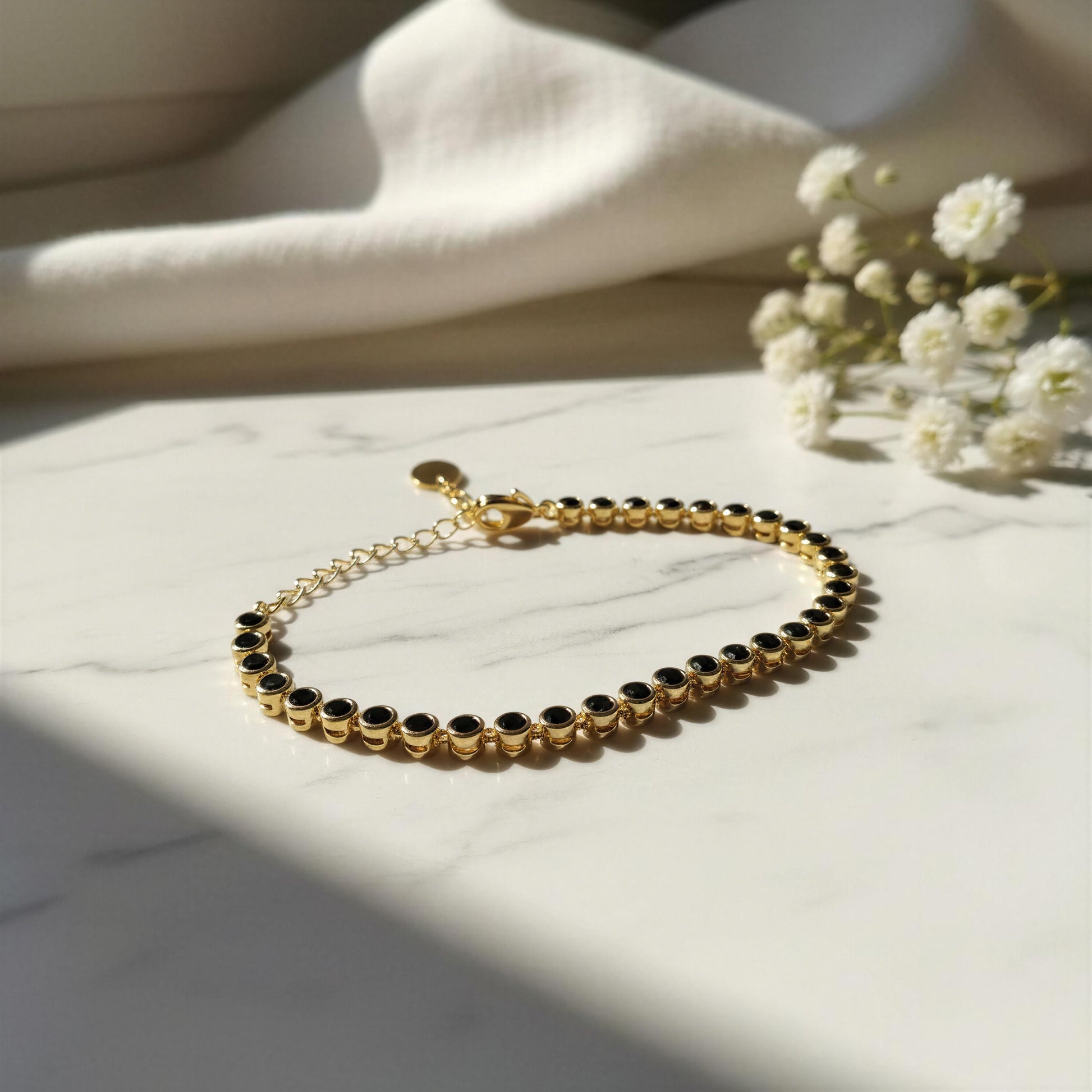 The Siena Black Tennis Bracelet from Chica Chica Jewelry, displayed on a white marble surface with soft shadows and white fabric flowers in the background.