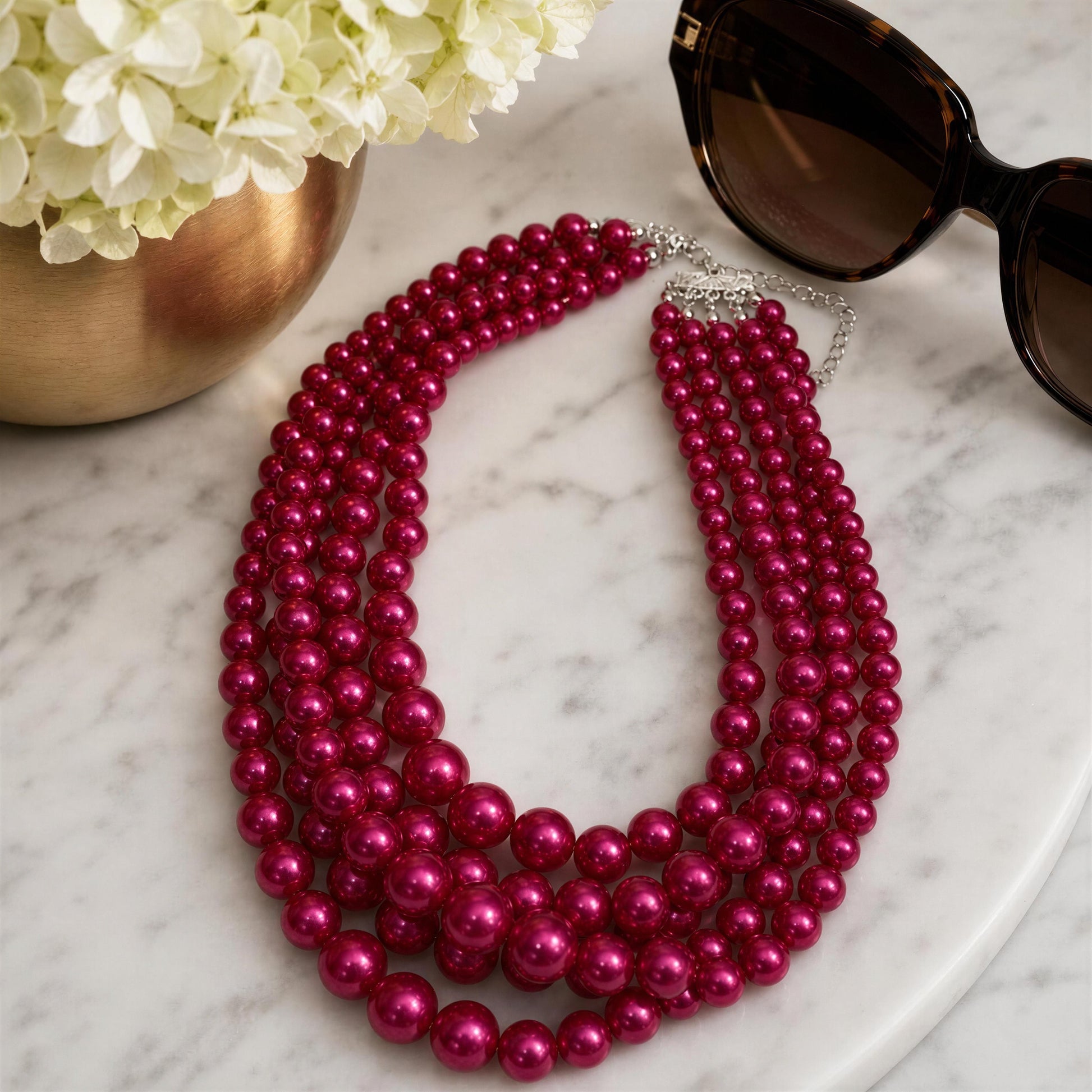 Pink pearl necklace on a marble surface with sunglasses and flowers.