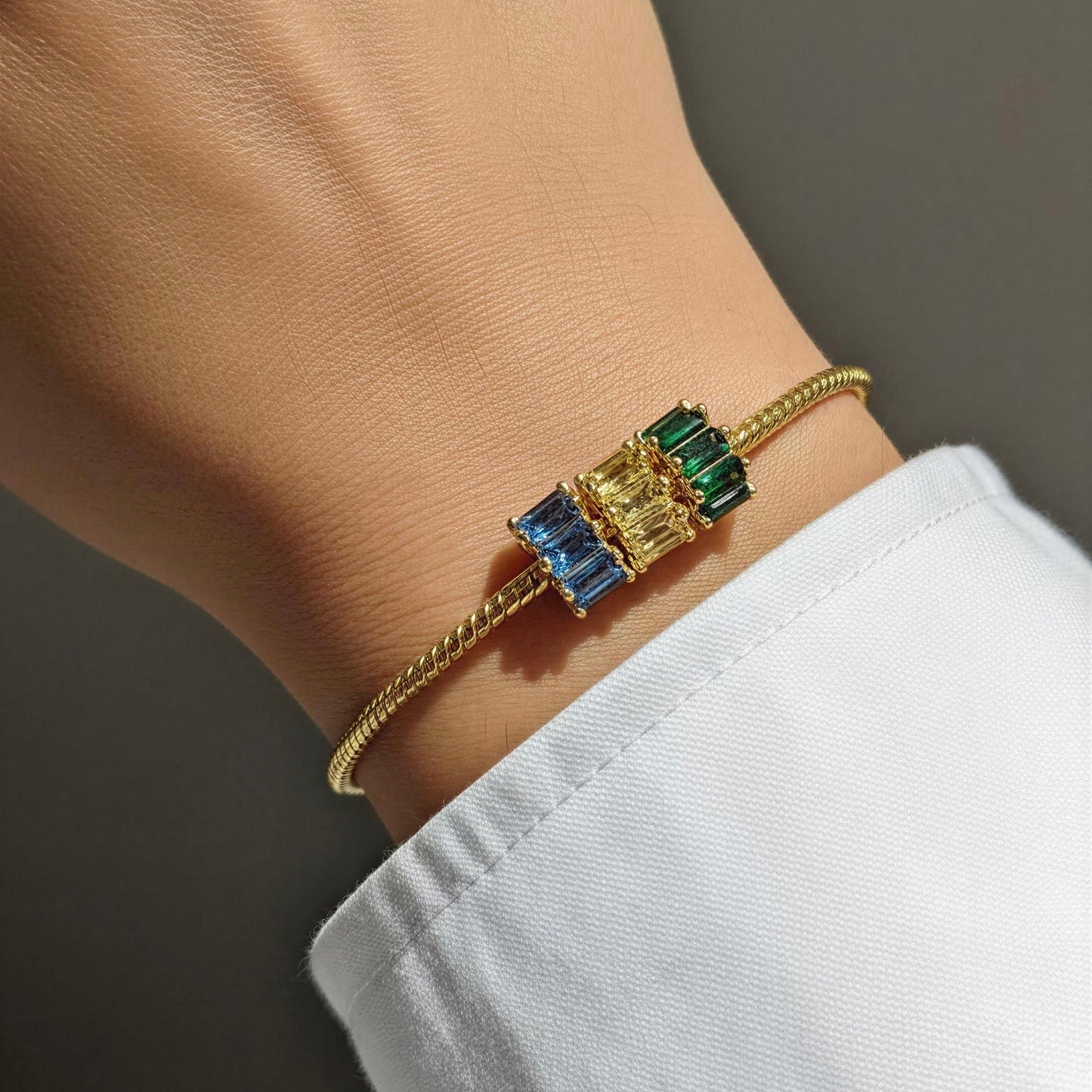 A gold rope bracelet with a central cluster of rectangular baguette charms in blue, yellow, and green, shown worn on a person's wrist with a white shirt cuff visible.