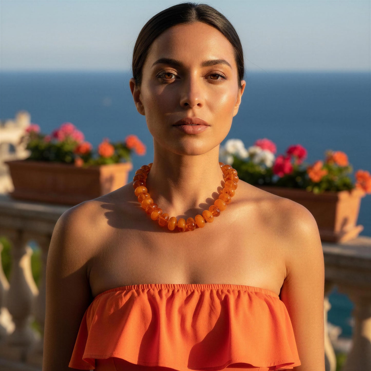 A woman with dark hair pulled back wears a chunky amber-orange beaded necklace and a matching orange strapless top, standing outdoors with a blurred ocean and flower boxes in the background.