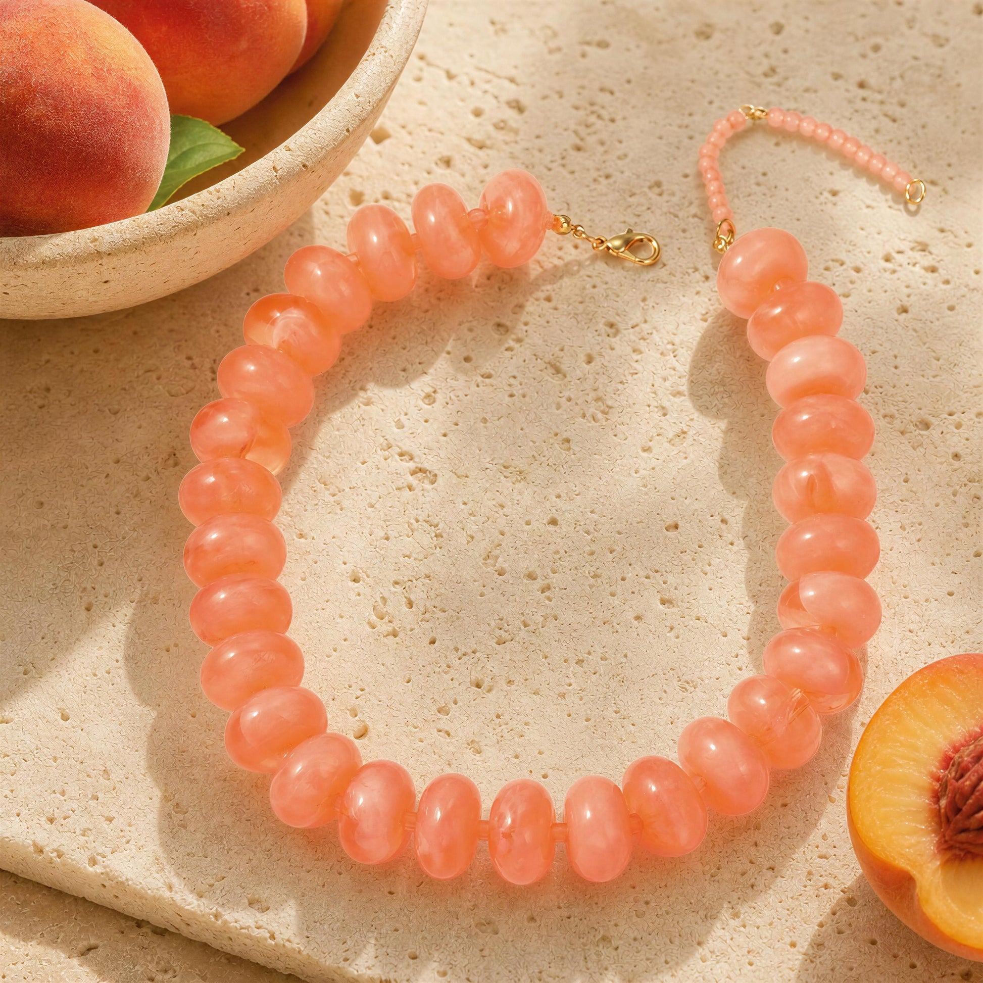 A peach-colored beaded necklace with a gold clasp, styled on a textured beige stone surface with peach slices and a bowl in the background.