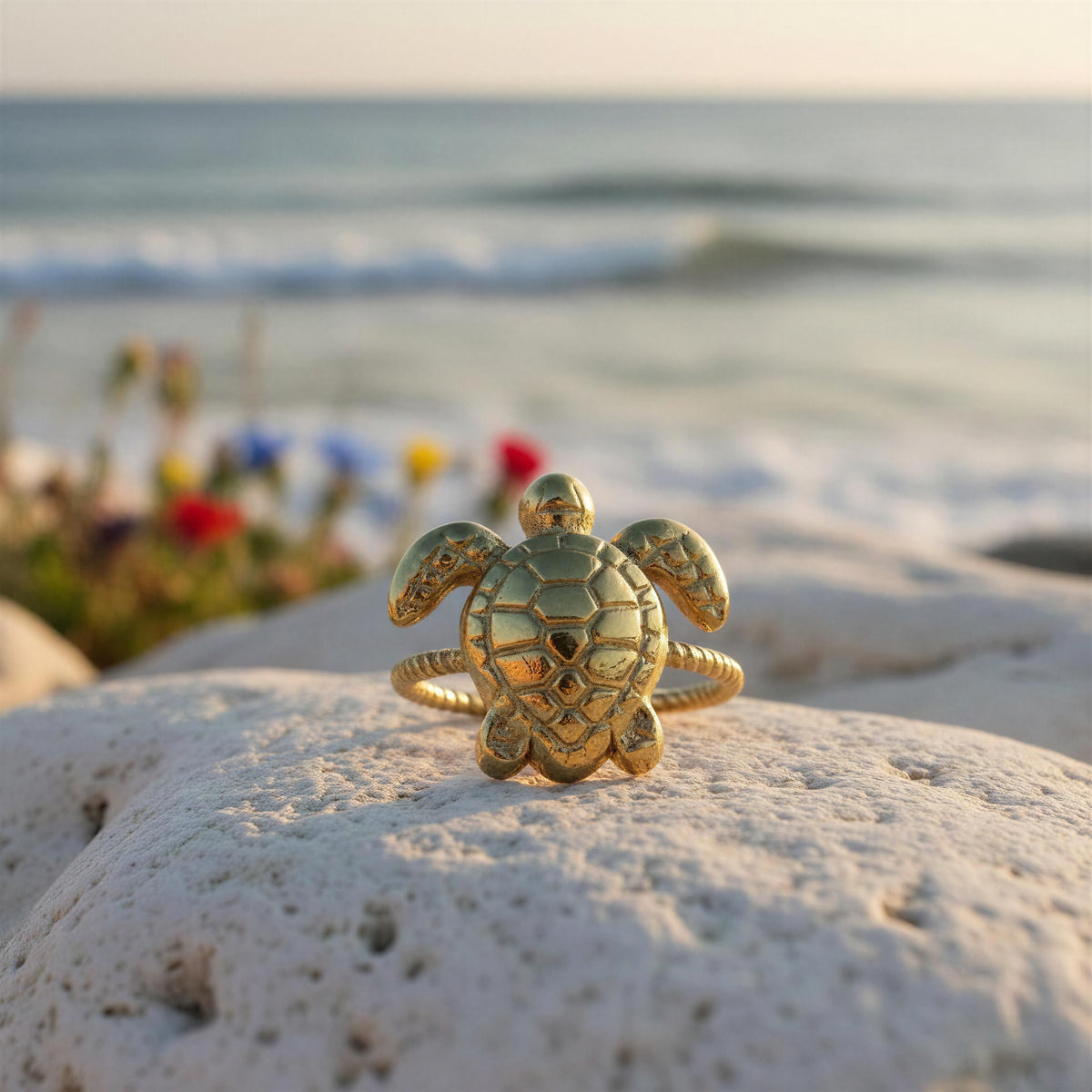 A gold sea turtle ring with a textured shell and band, resting on a white rock with a blurred beach and ocean background.