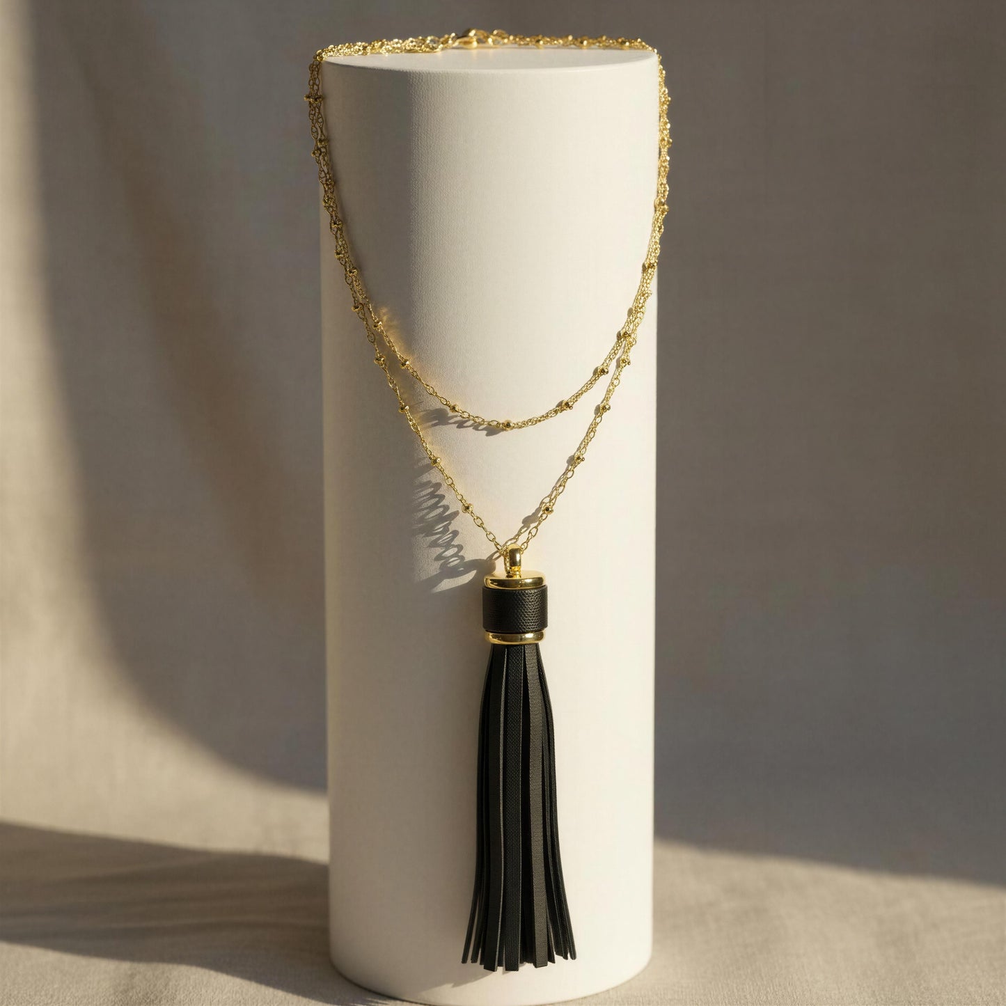 The Frangia Ebano Necklace with a black tassel pendant, displayed on a white cylindrical stand against a neutral beige background.