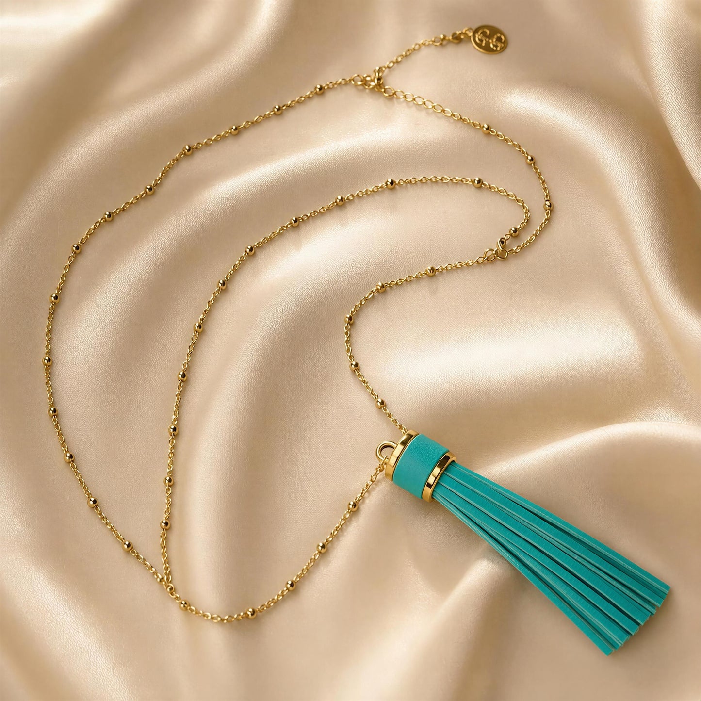 The Frangia Firuze Necklace, a gold chain necklace with a turquoise tassel pendant, styled on a draped beige satin fabric background.