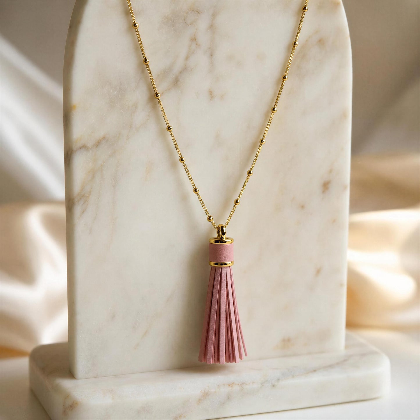 The Frangia Rosa Necklace, an 18k gold plated  sattelite beaded chain necklace with a pink tassel pendant, displayed on a white marble stand against a soft cream satin background. 