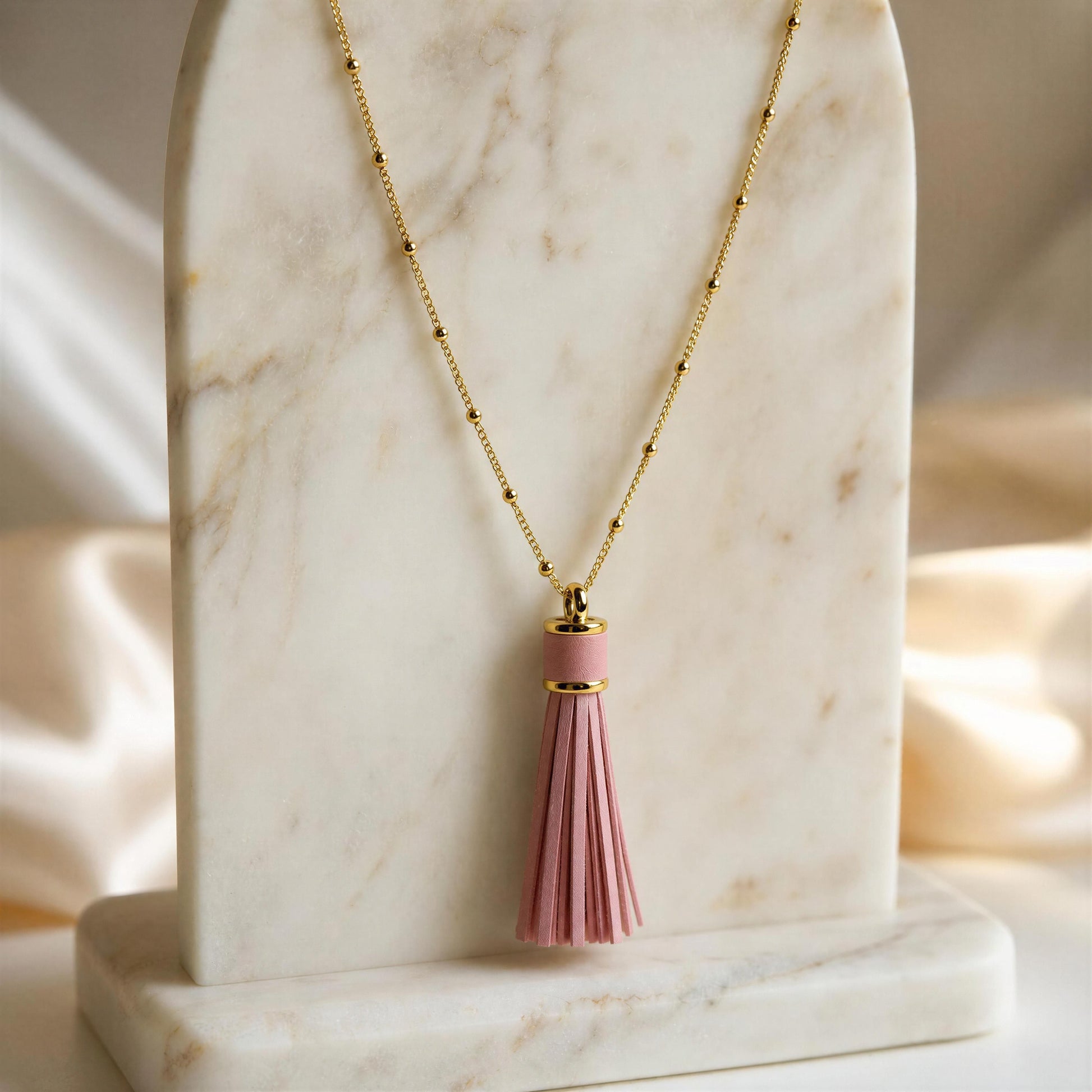 The Frangia Rosa Necklace, an 18k gold plated  sattelite beaded chain necklace with a pink tassel pendant, displayed on a white marble stand against a soft cream satin background. 