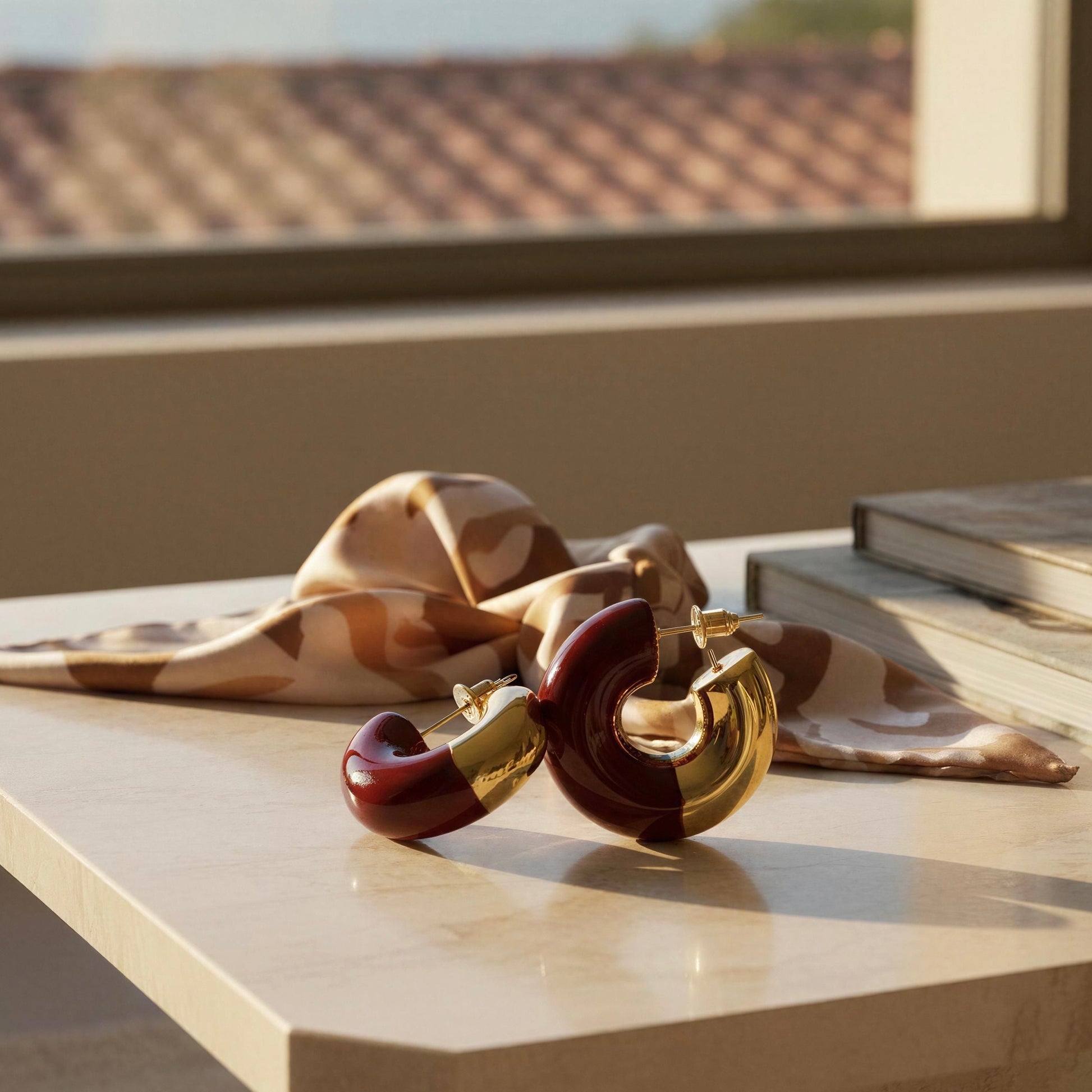 A pair of chunky gold-tone hoop earrings with deep burgundy red inner sections, styled on a light marble tabletop next to a patterned silk scarf and books.