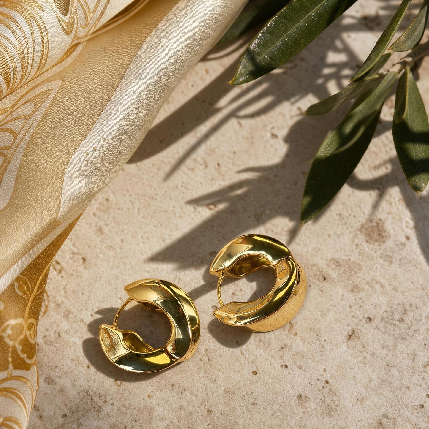 A pair of gold crescent-shaped hoop earrings with a polished, reflective finish, styled on a textured beige stone surface next to a gold patterned silk scarf and green leaves.