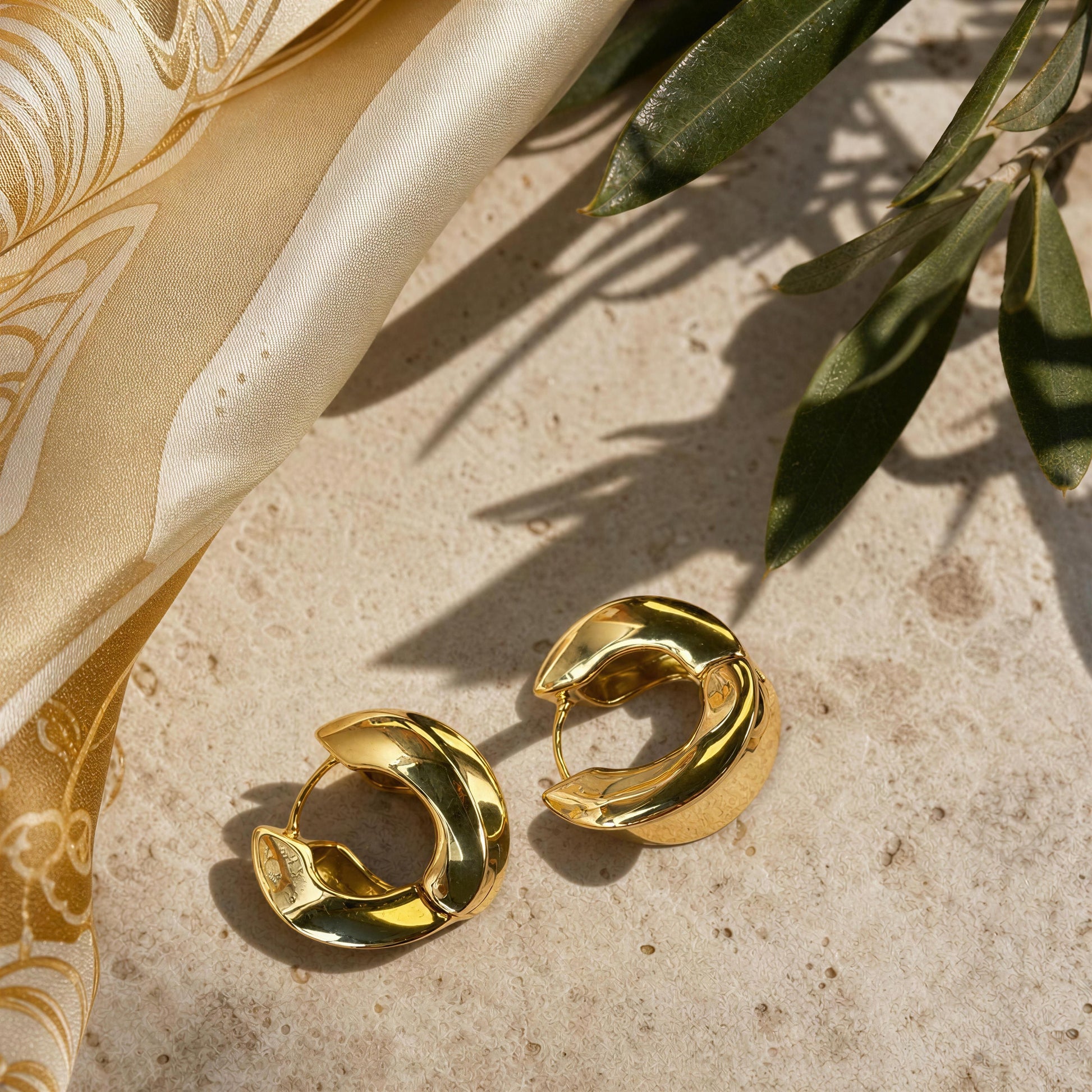 A pair of gold crescent-shaped hoop earrings with a polished, reflective finish, styled on a textured beige stone surface next to a gold patterned silk scarf and green leaves.
