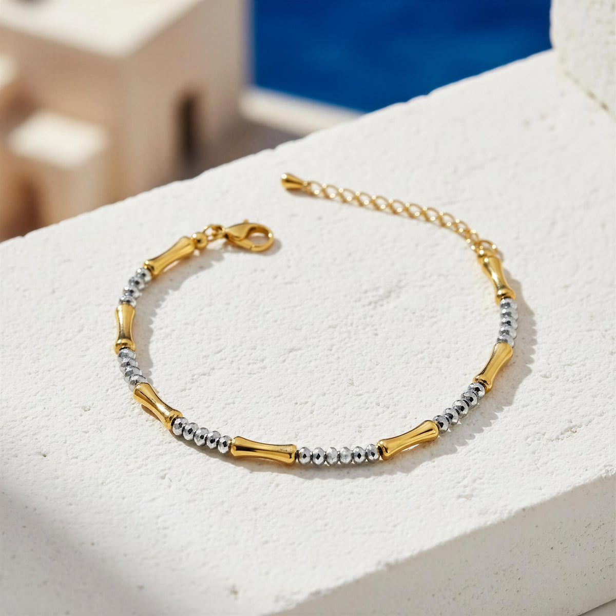 A gold and silver beaded bracelet with bamboo-shaped gold links, displayed on a textured white stone surface with a blurred blue background.