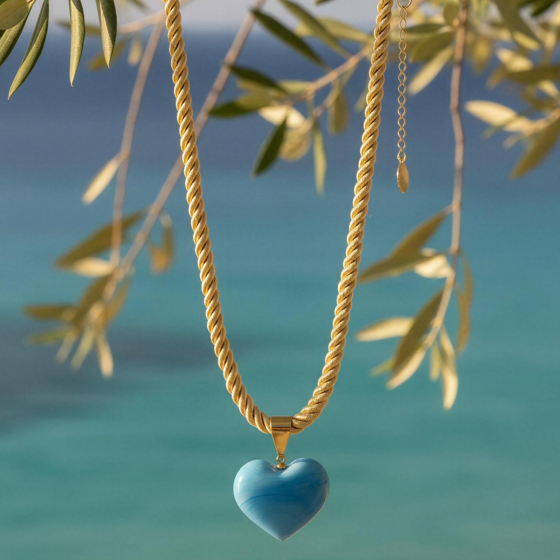The Mavi Murano-style blue glass heart necklace with a soft braided cord and 18k gold-plated details by Chica Chica Jewelry at coastline.