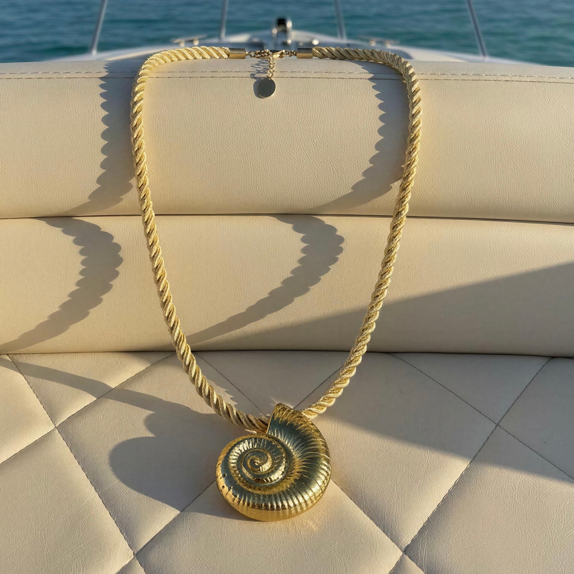 A cream rope-chain necklace with a  shell pendant, displayed on a cream-colored quilted boat seat with the ocean in the background.
