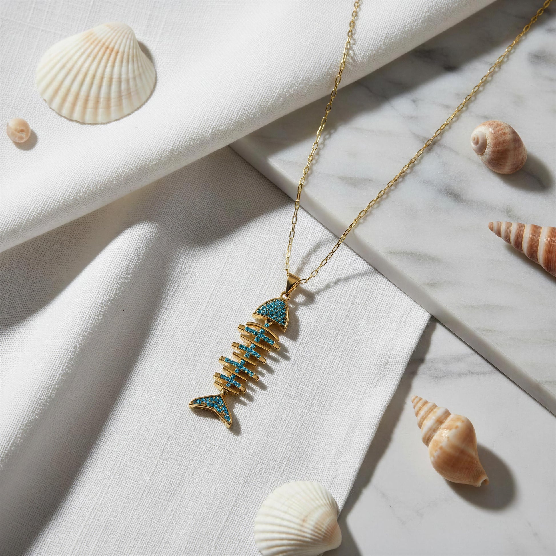 A gold fishbone pendant necklace with turquoise blue inlay, styled on a white draped fabric and marble slab with seashells.