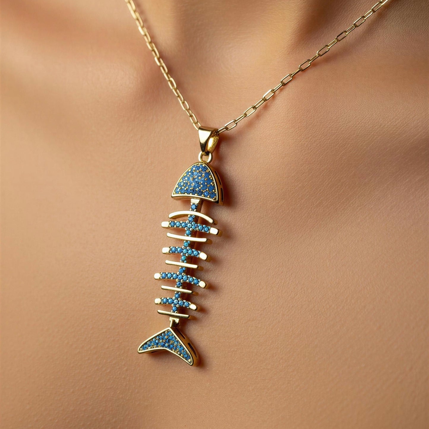 An 18k gold plated fishbone pendant necklace with blue stone accents, shown worn on a model's neck against a warm beige skin background.