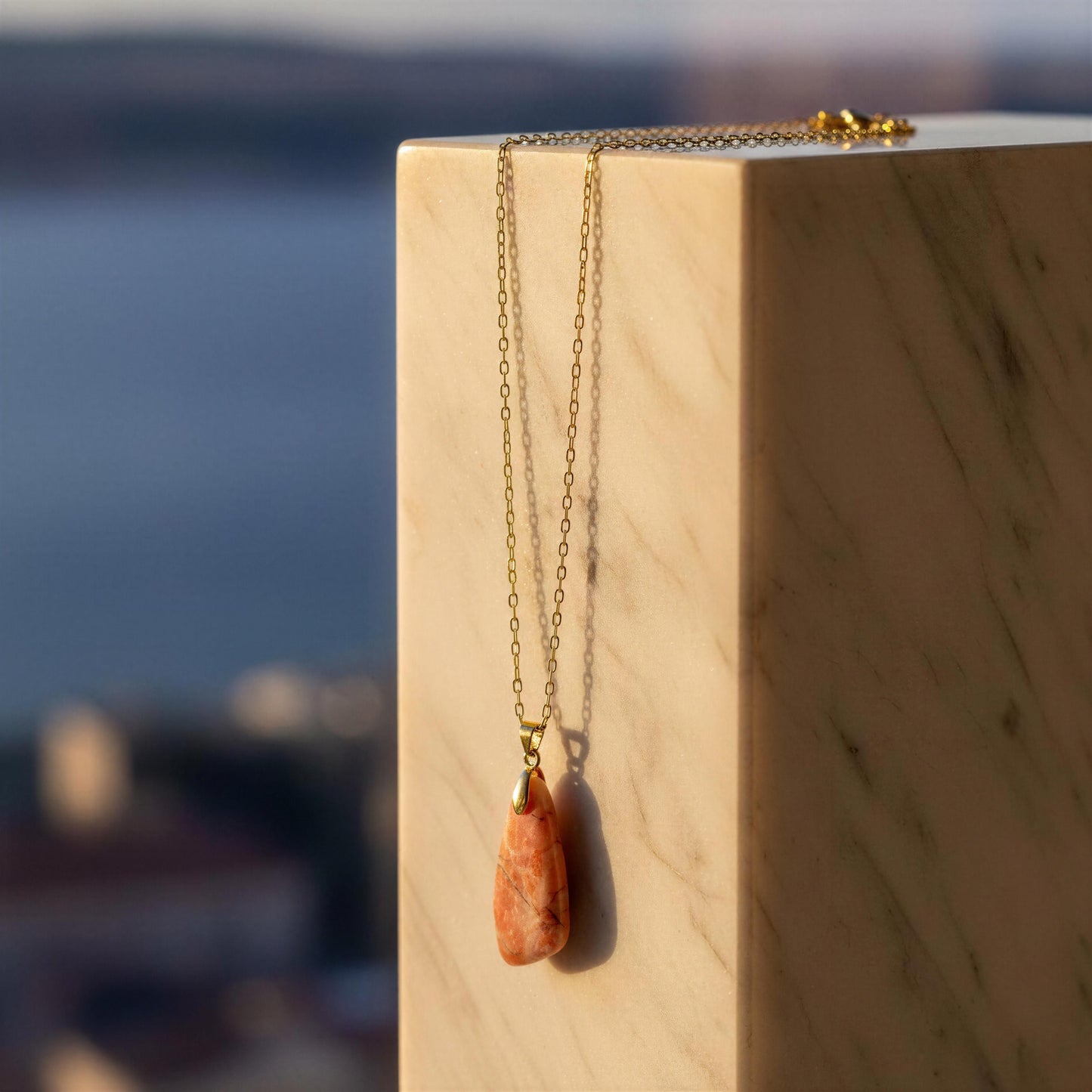 A gold chain necklace with a teardrop-shaped coral orange stone pendant, draped over a light beige marble block with a blurred outdoor background.