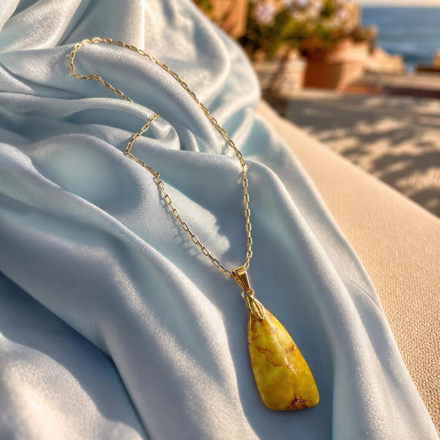A gold chain necklace with a yellow marbled stone pendant draped over light blue satin fabric, with a blurred outdoor background.