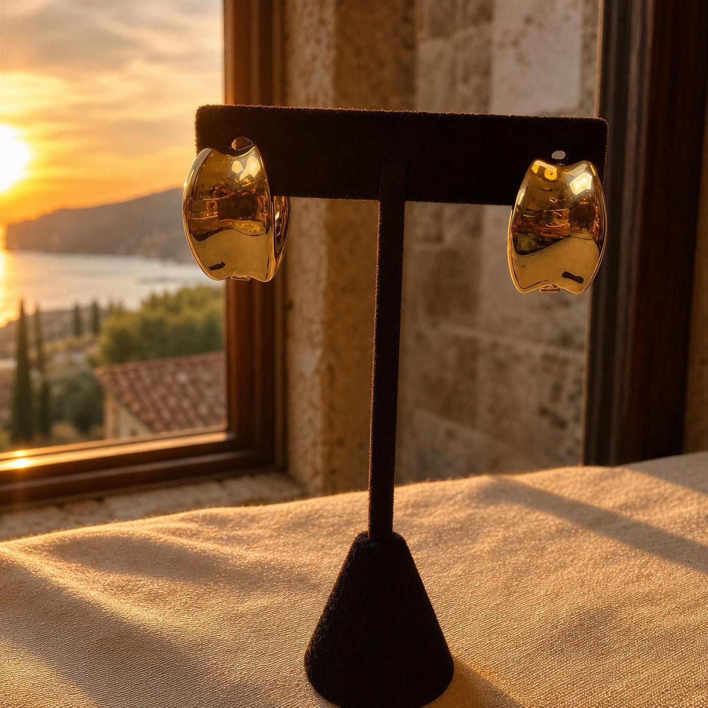 A pair of chunky, organic-shaped gold hoop earrings displayed on a black velvet jewelry stand, set against a warm sunset window view of a coastal town.