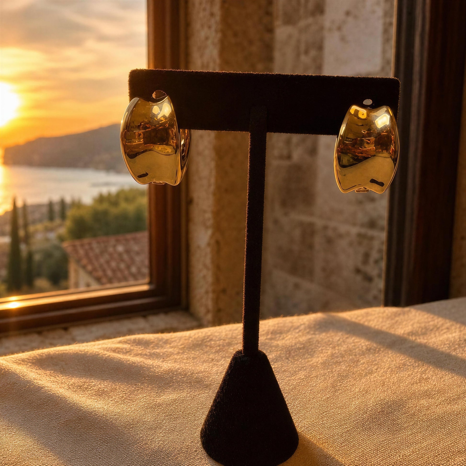 A pair of chunky, organic-shaped gold hoop earrings displayed on a black velvet jewelry stand, set against a warm sunset window view of a coastal town.