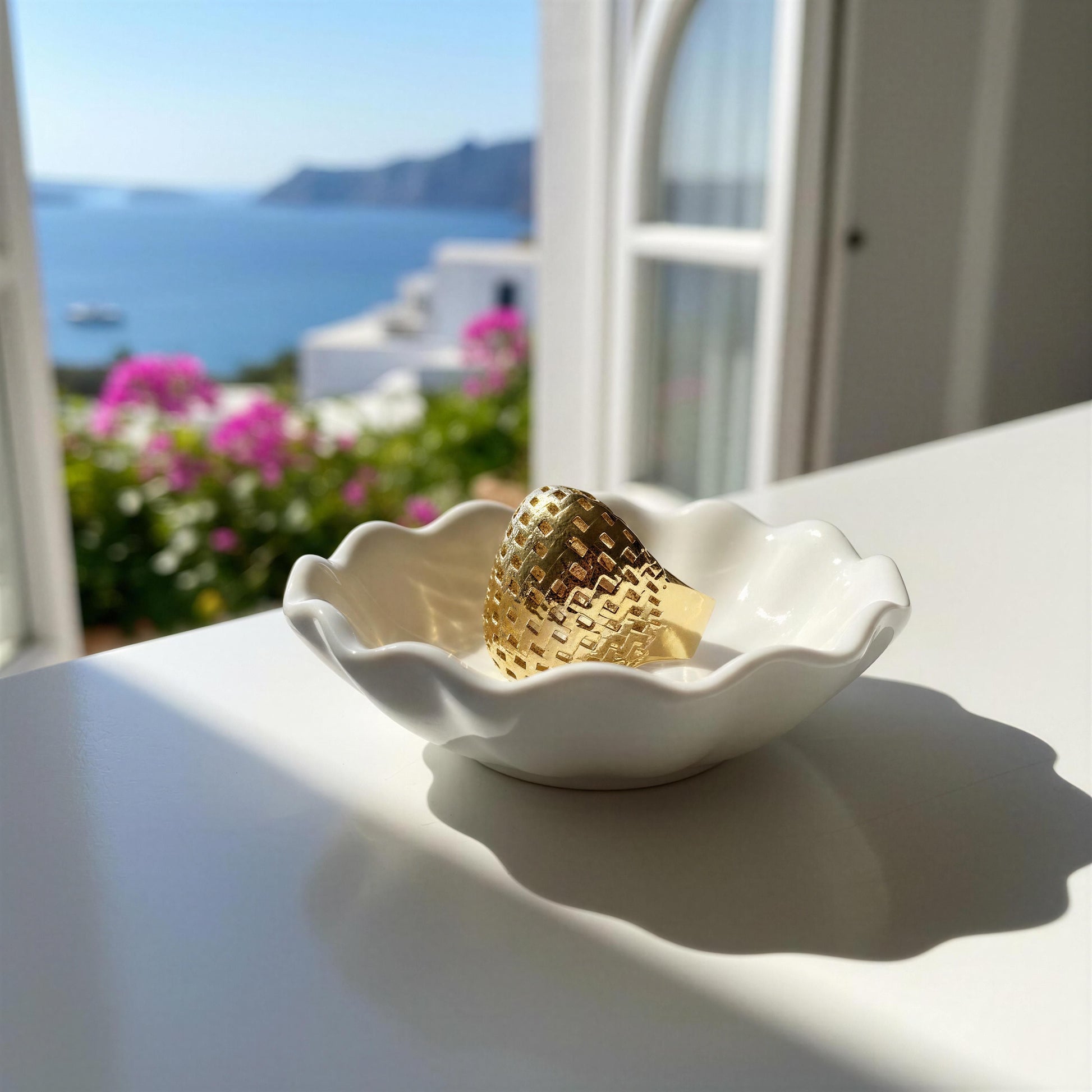 The Trama Ring, a textured gold ring displayed in a white ceramic dish on a white table, with a blurred Mediterranean coastal background featuring blue sea, white buildings, and pink flowers.