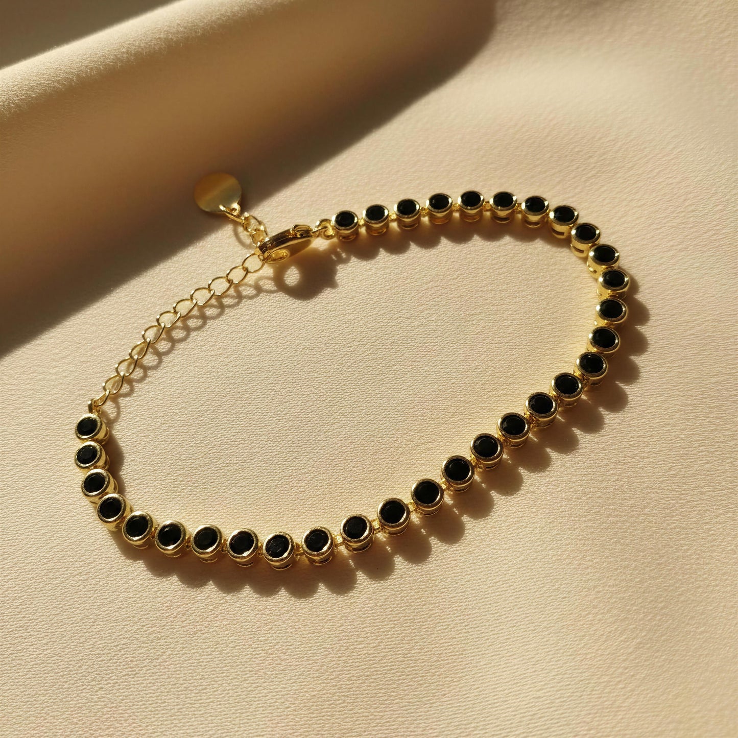 The Siena Black Tennis Bracelet from Chica Chica Jewelry displayed on a textured beige surface with warm sunlight and shadows.
