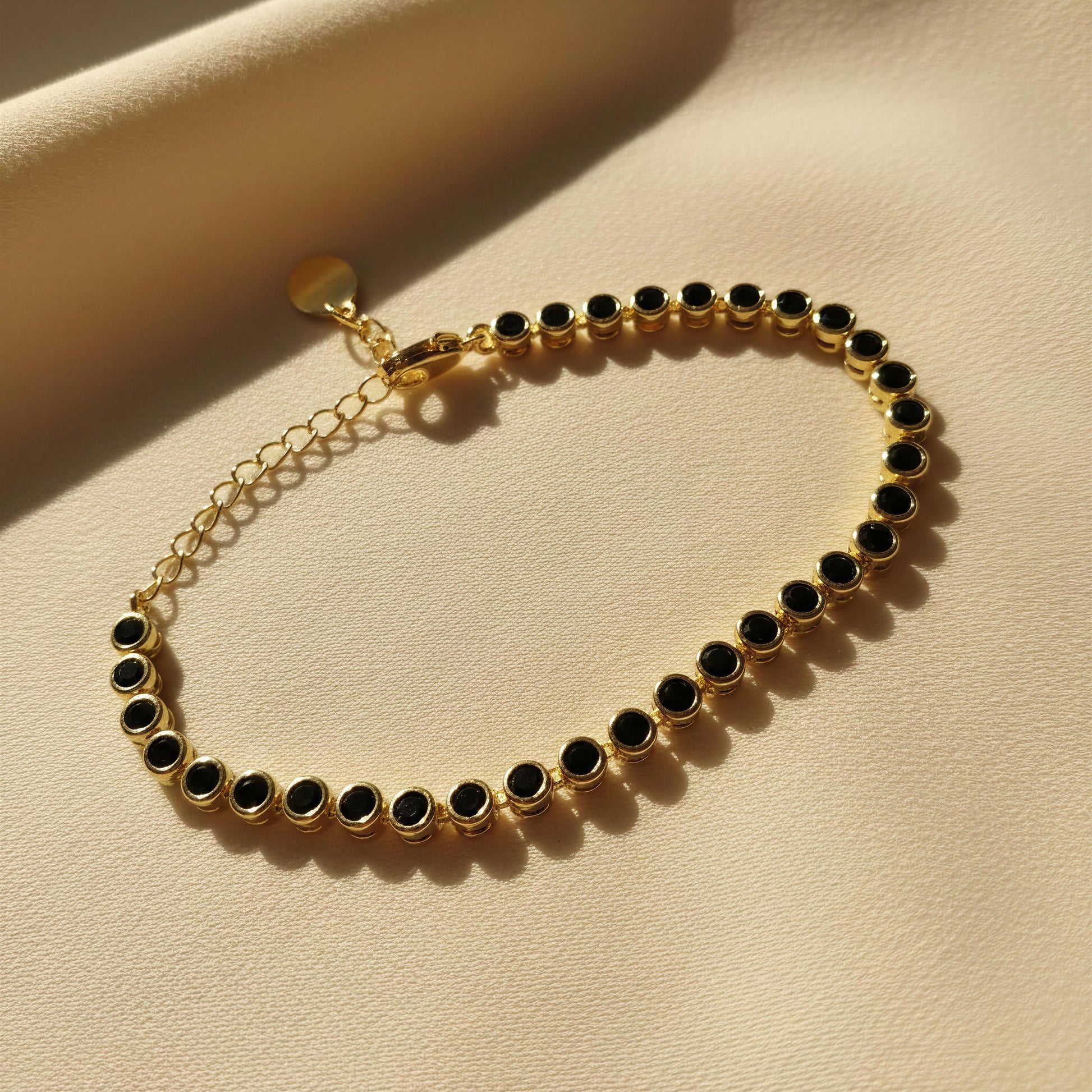 The Siena Black Tennis Bracelet from Chica Chica Jewelry displayed on a textured beige surface with warm sunlight and shadows.