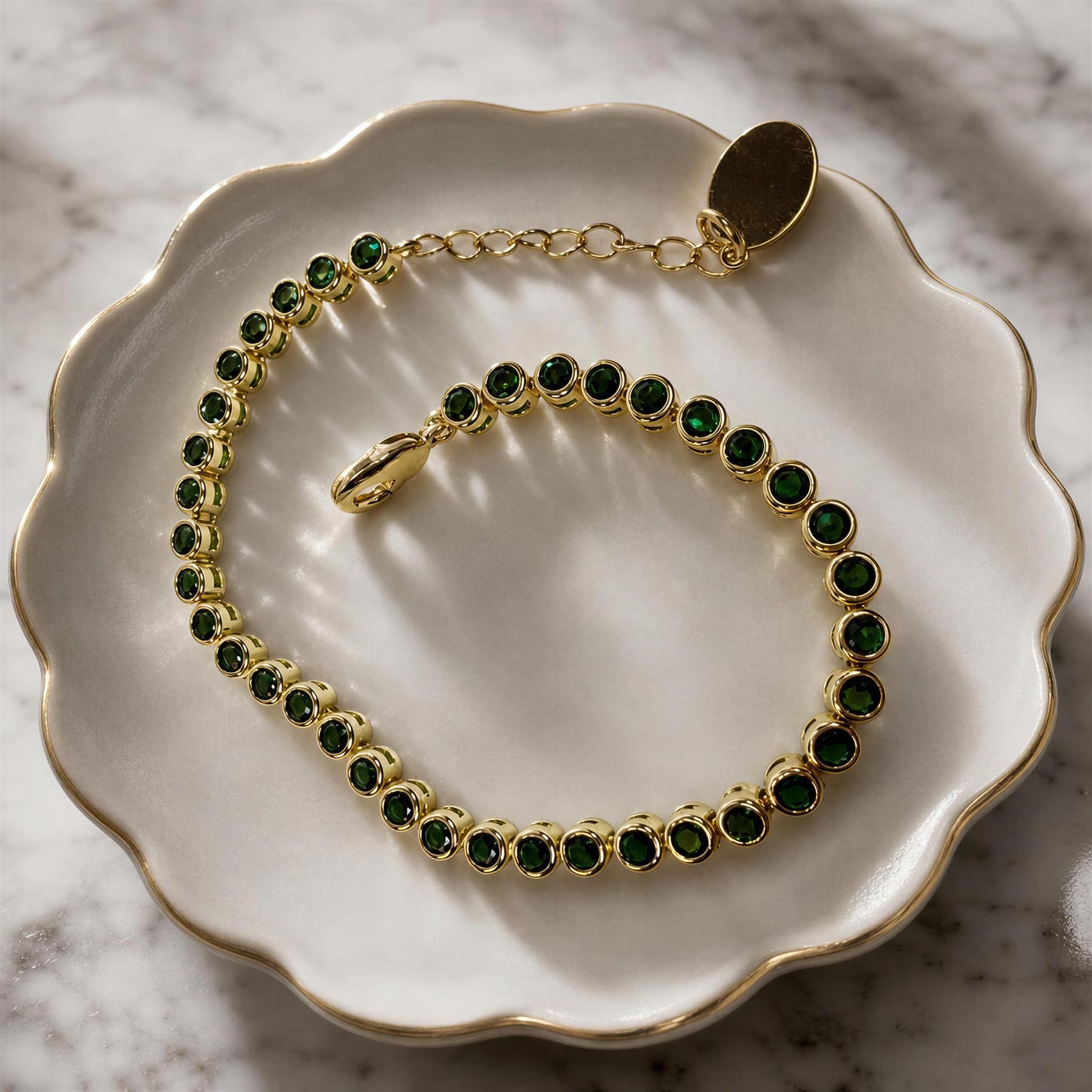 The Siena Emerald Tennis Bracelet with bezel-set emerald green cubic zirconia in 18K gold-plated brass. displayed on a white scalloped dish over a marble surface.