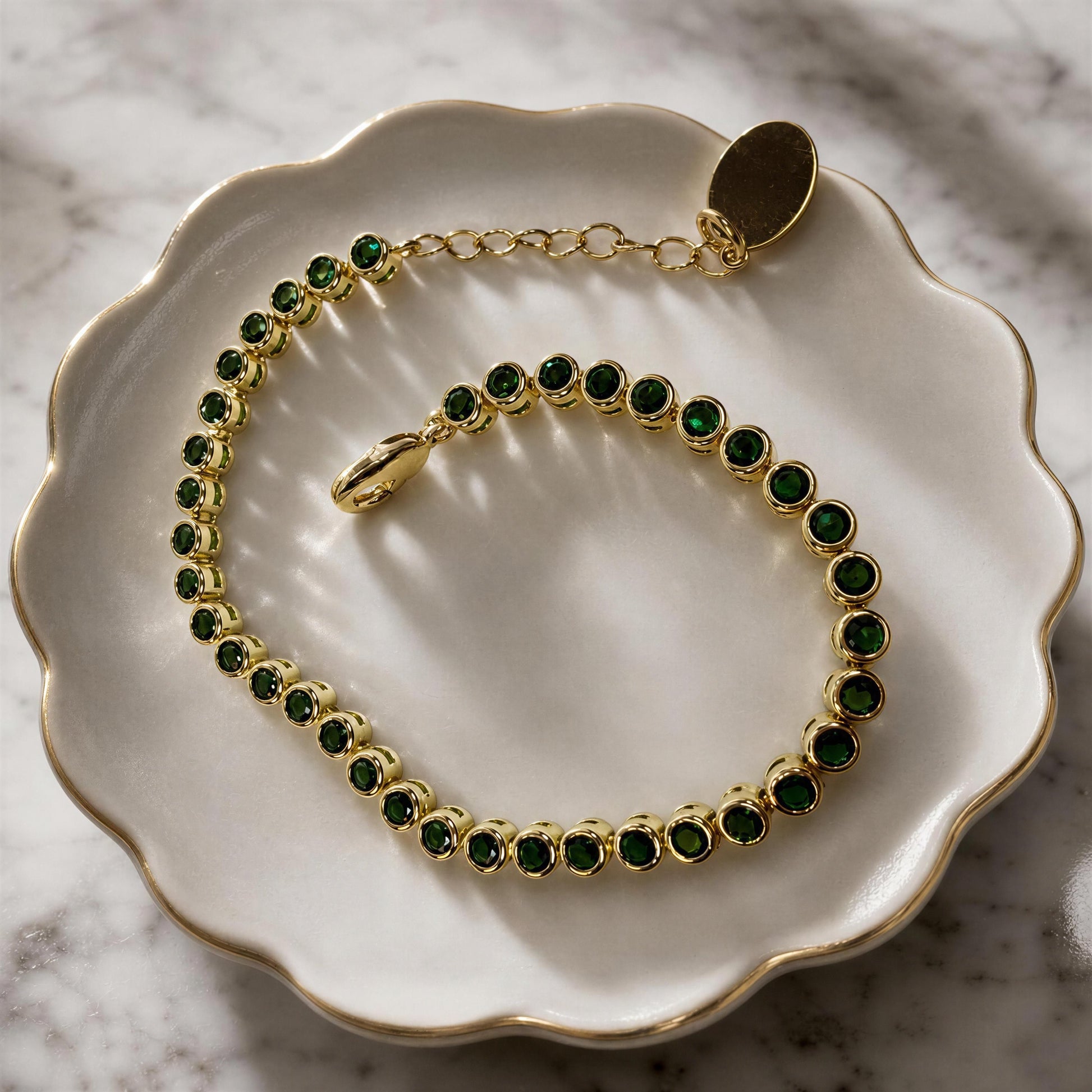 The Siena Emerald Tennis Bracelet with bezel-set emerald green cubic zirconia in 18K gold-plated brass. displayed on a white scalloped dish over a marble surface.