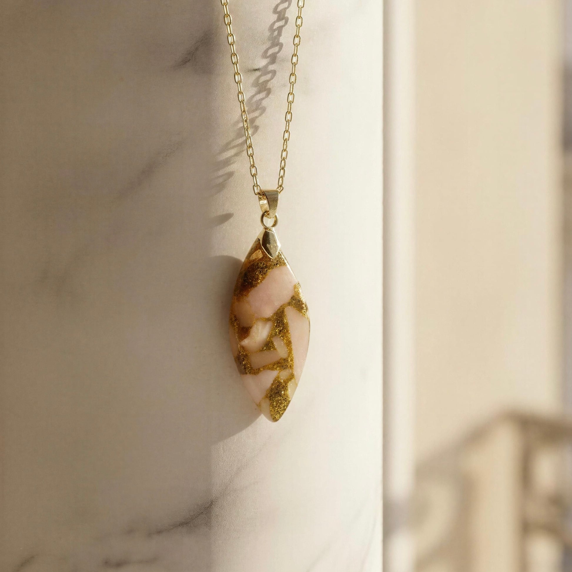 The Pietra Cipria Natural Stone Necklace, a  gold paperclip chain necklace with a teardrop-shaped pendant made of pale pink stone with gold glitter inclusions, resting against a white marble column.