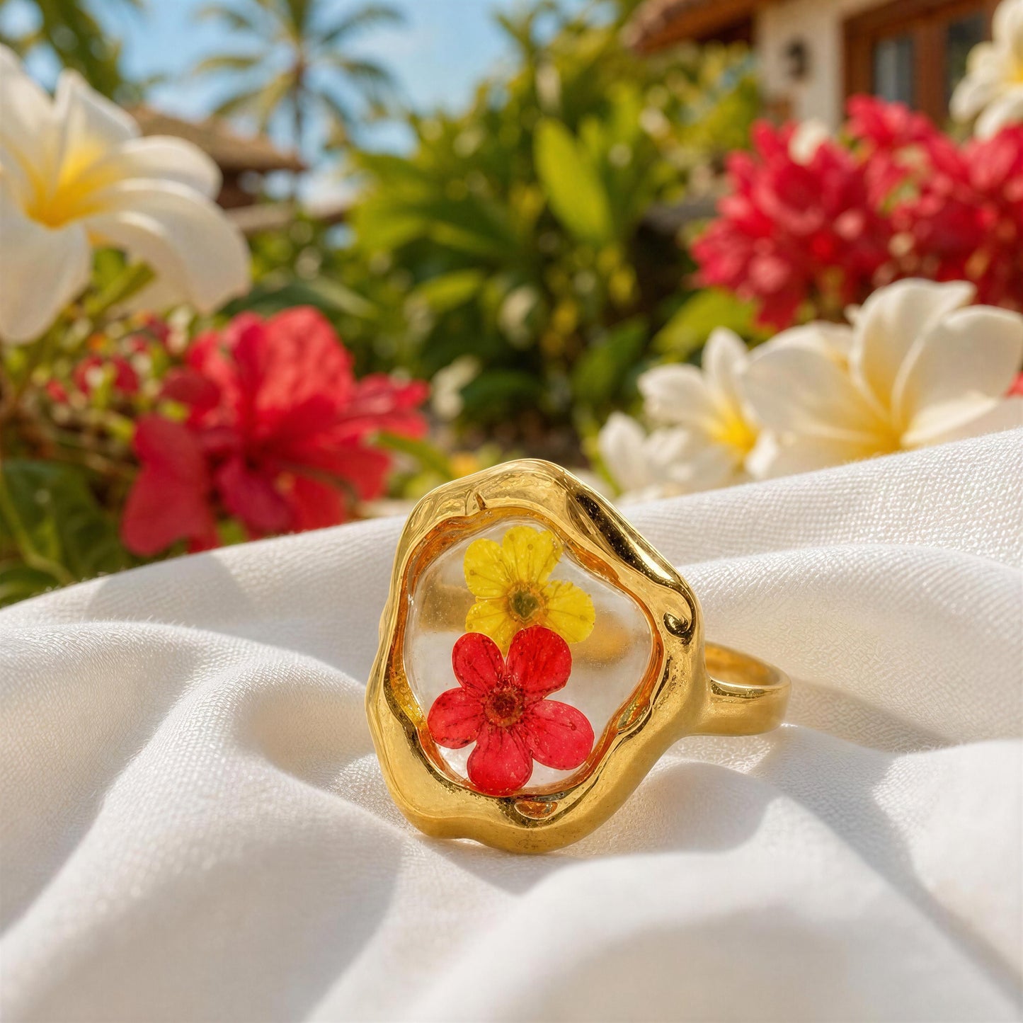 Gold ring with embedded red and yellow flowers on a white fabric background with tropical flowers in the background.