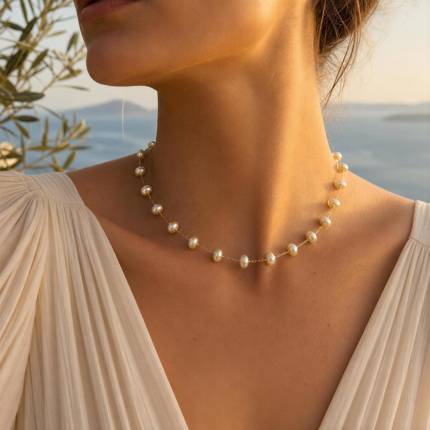 A close-up lifestyle shot of a woman wearing the Lunaria_Pearl_Necklace a delicate gold chain necklace with small cream donut freshwater pearls, set against a warm, sunlit background with a blurred sea and olive tree.