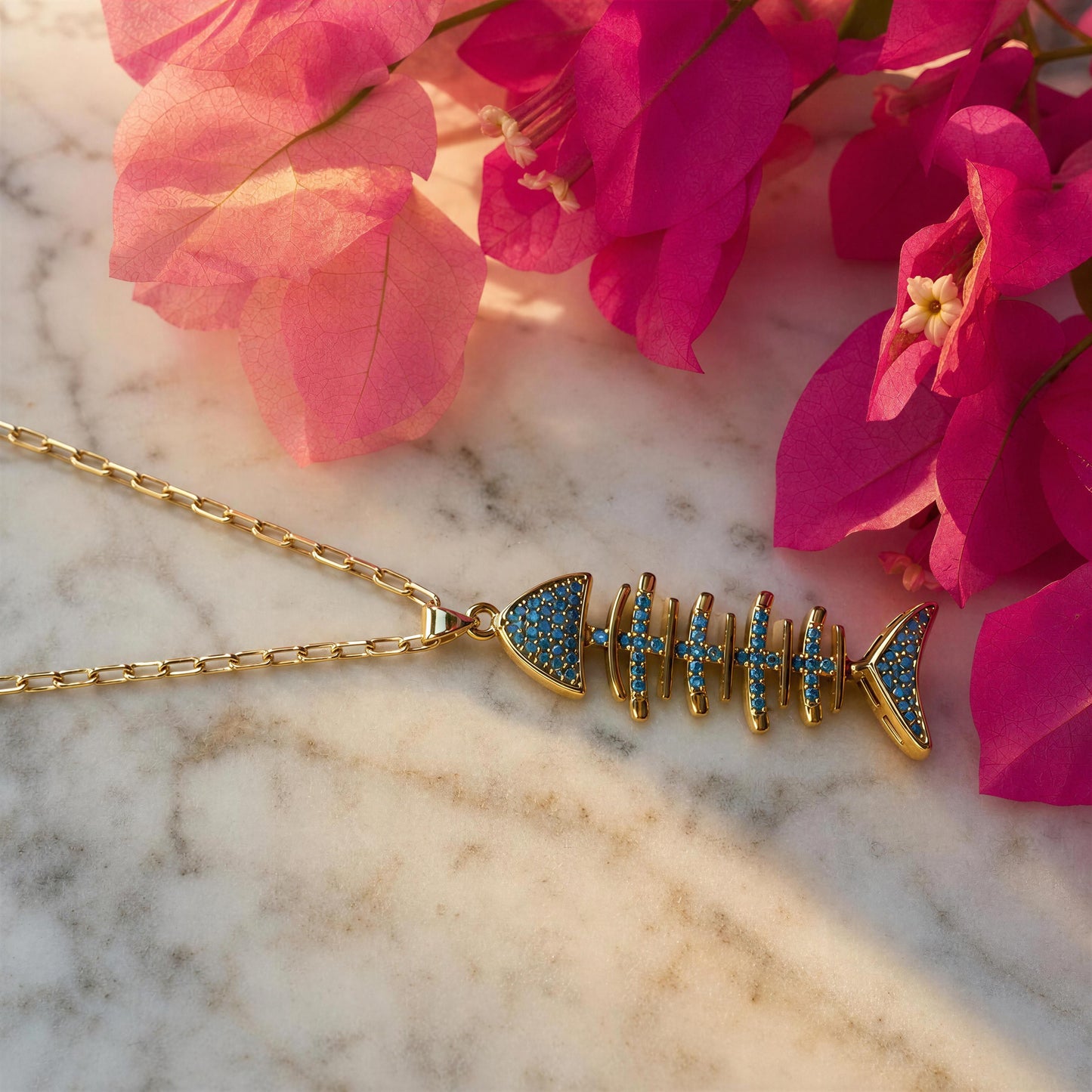 Gold fish skeleton pendant necklace with blue pavé accents, displayed on a white marble surface with pink flowers in the background.