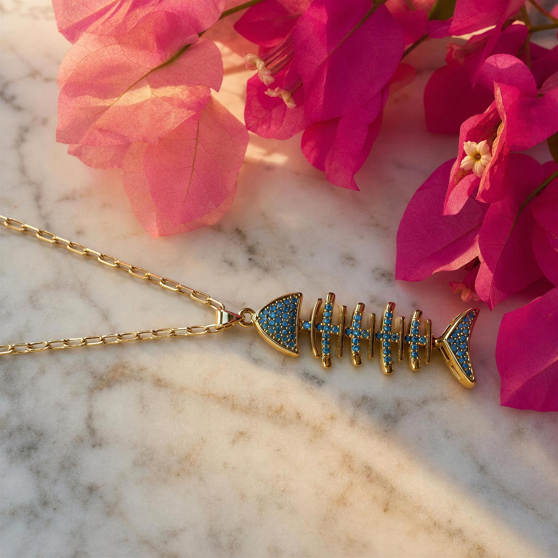 Gold fish skeleton pendant necklace with blue pavé accents, displayed on a white marble surface with pink flowers in the background.