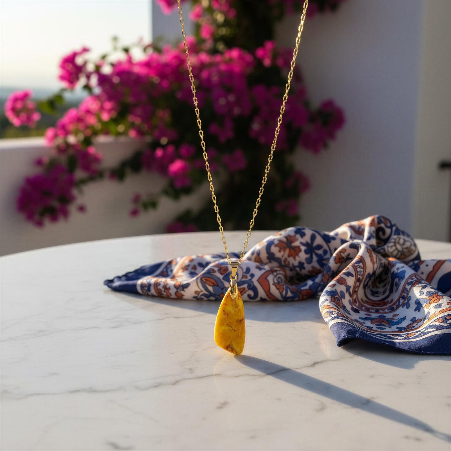 The Pietra Sole Natural Stone Necklace, a gold paperclip chain necklace with a yellow teardrop pendant resting on a white marble table, styled with a blue and orange paisley patterned scarf, with pink flowers in the background.