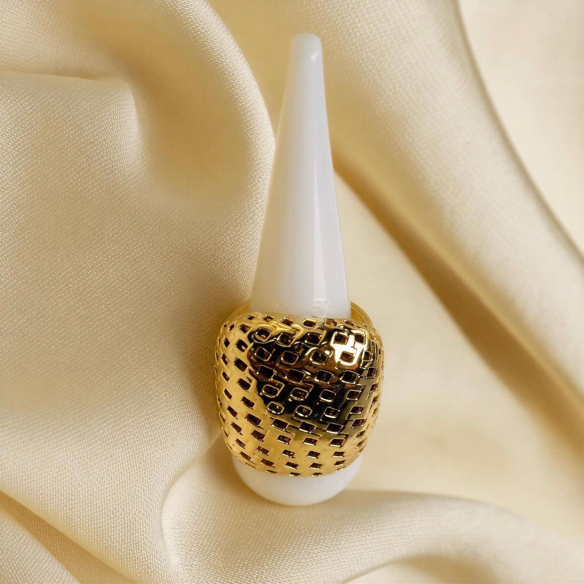 A gold ring with a textured, openwork geometric pattern, displayed on a white cone stand against a draped cream satin fabric background.